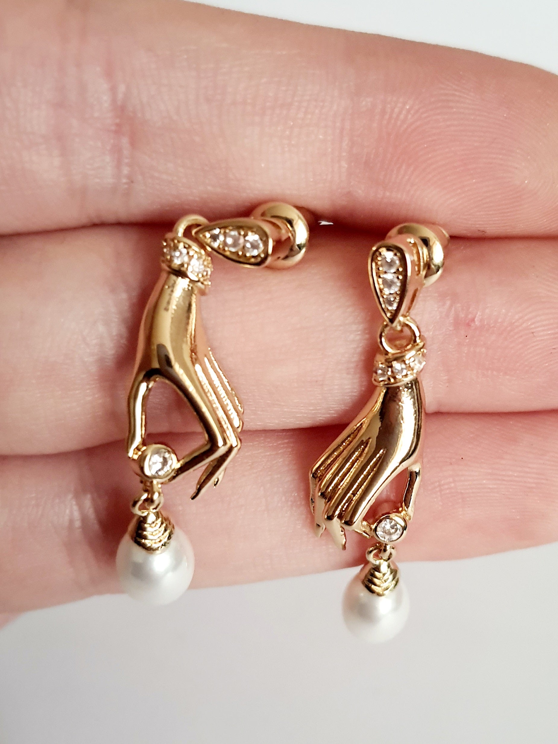 Kolczyki złote ze stali nierdzewnej w kształcie dłoni trzymających perłę, zdobione cyrkoniami, długość 4 cm, artystyczny styl glamour i elegancja. Rouge Bizu.
