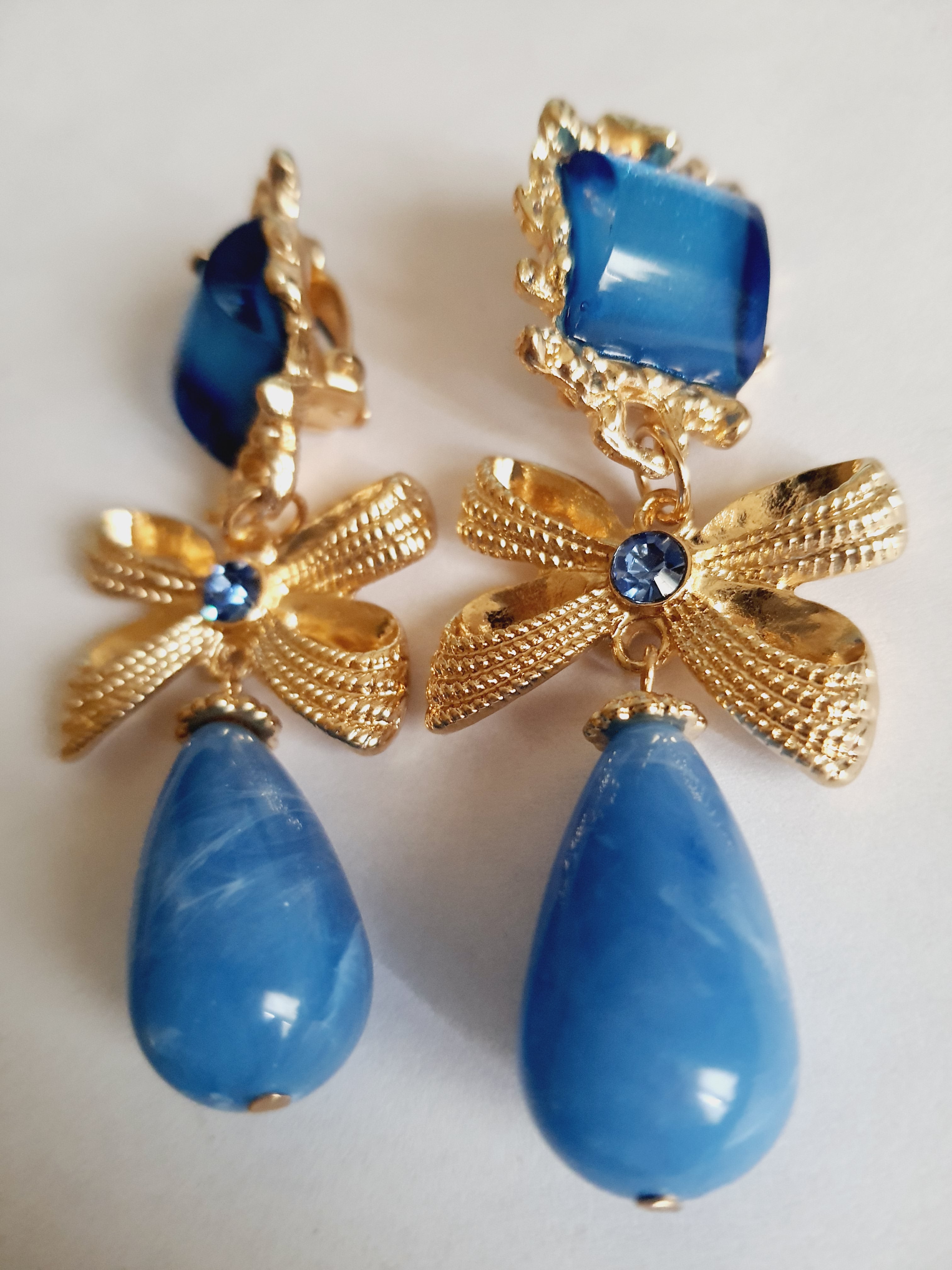 Złote klipsy ze stali nierdzewnej z niebieskimi kryształami. Dekoracyjne złote kokardy z błękitnym kamieniem i dużą kroplą w odcieniu niebieskiego. Styl glamour, romantyczna elegancja. Długość 7 cm. Rouge Bizu.