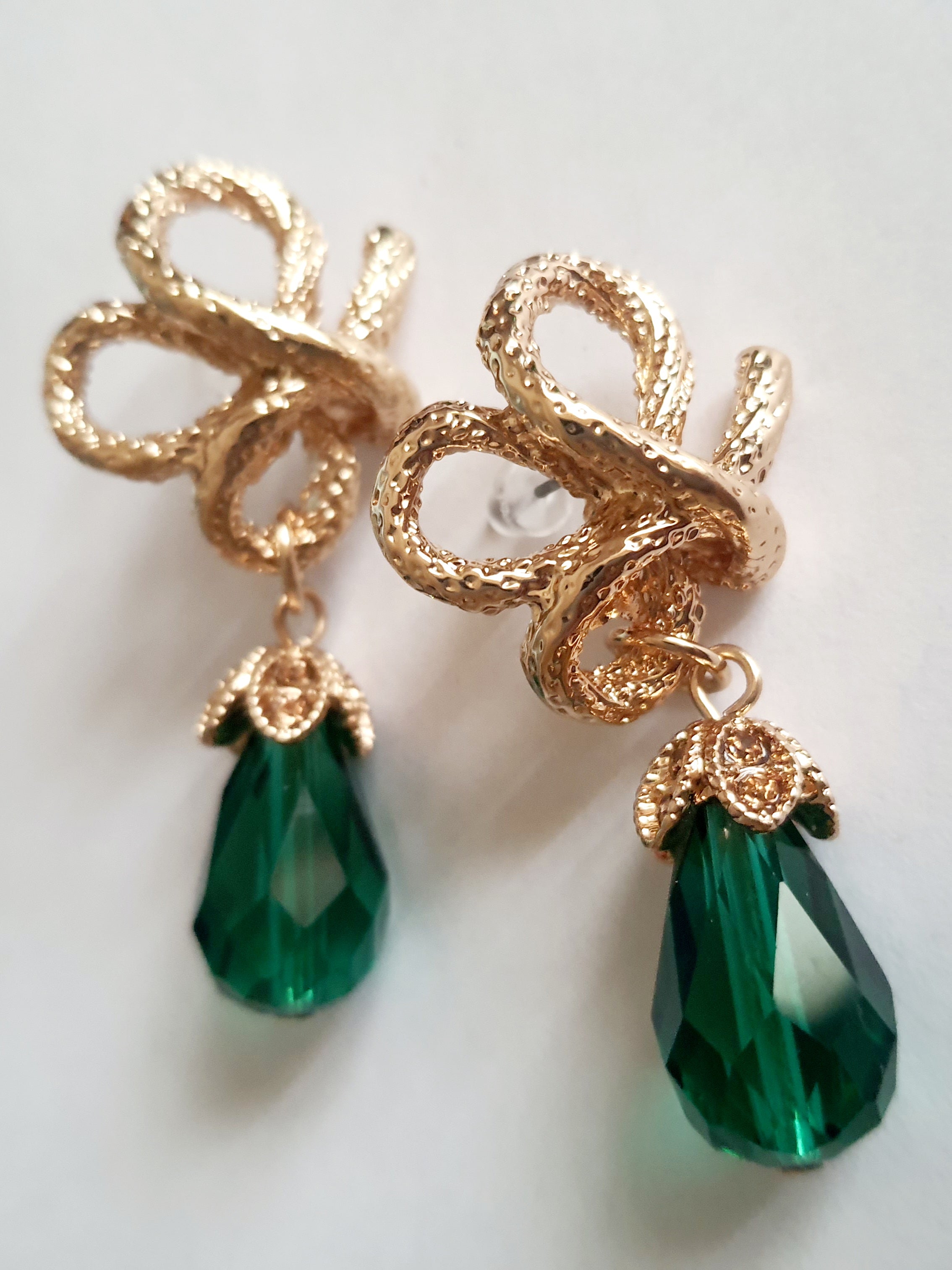 Złote kolczyki ze stali nierdzewnej w stylu glamour, ozdobione rzeźbioną kokardą i zielonym kryształem w kształcie kropli. Elegancja i szmaragdowy blask. Długość 6 cm. Rouge Bizu.