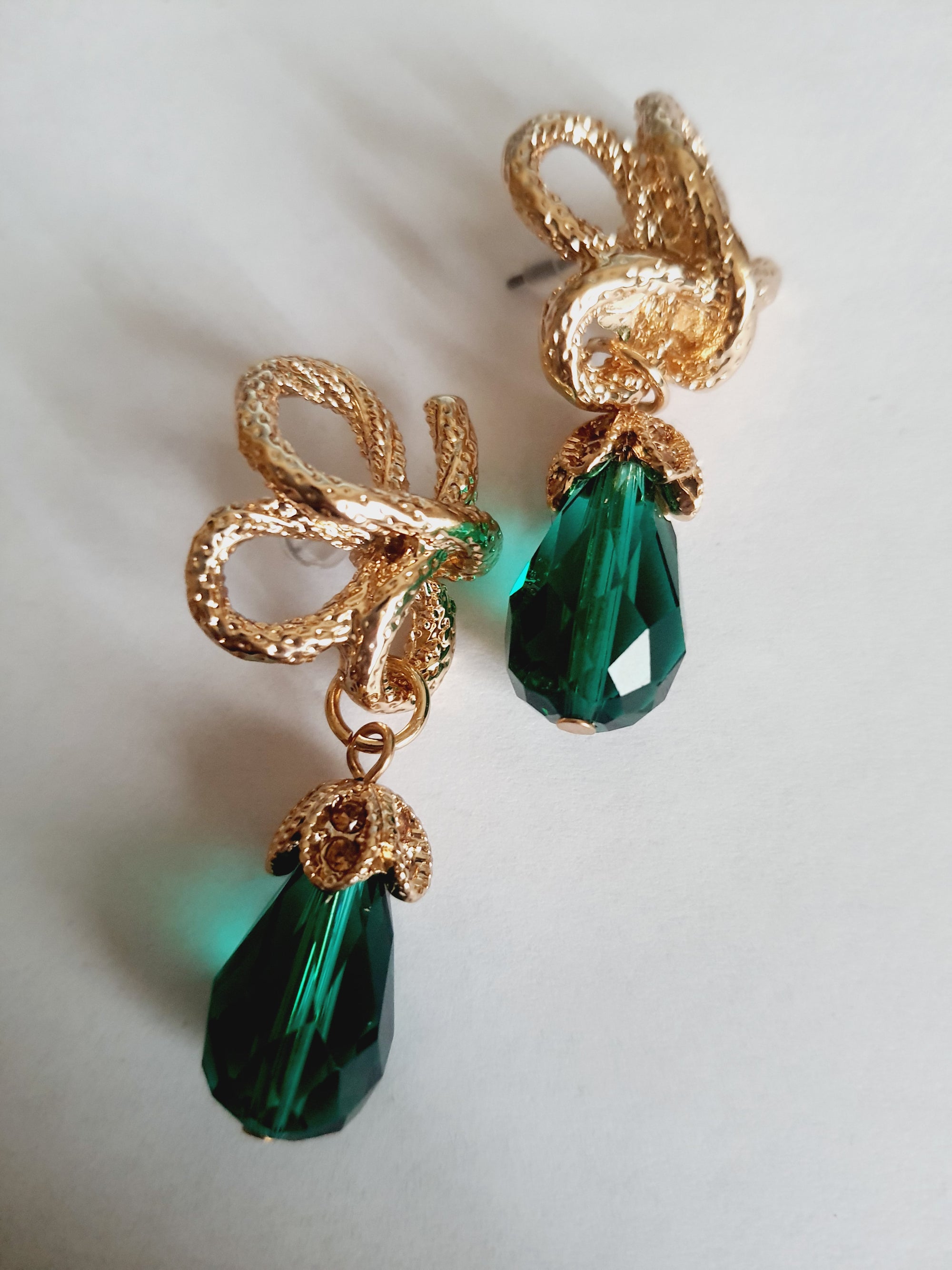 Złote kolczyki ze stali nierdzewnej w stylu glamour, ozdobione rzeźbioną kokardą i zielonym kryształem w kształcie kropli. Elegancja i szmaragdowy blask. Długość 6 cm. Rouge Bizu.