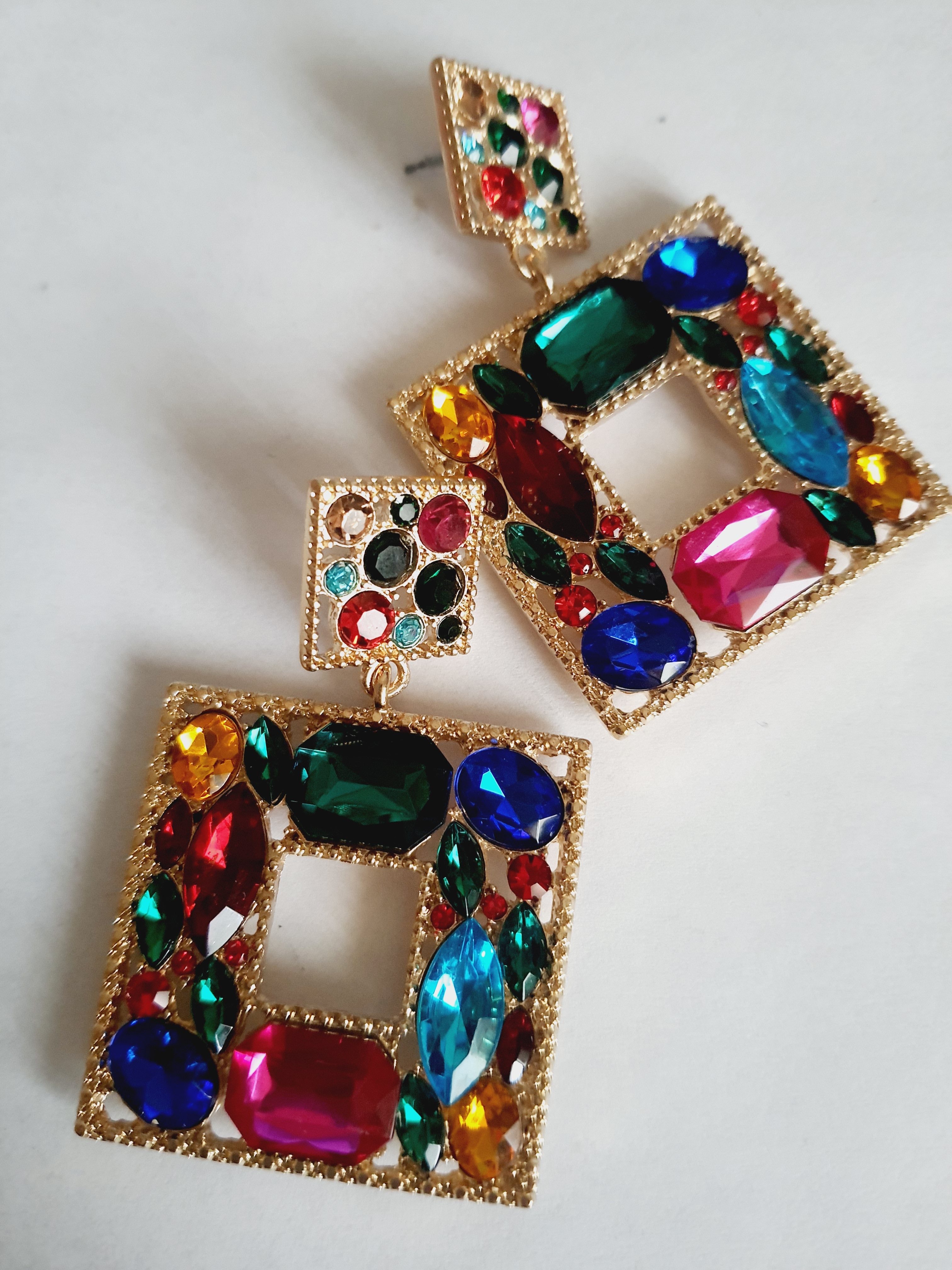 Złote kolczyki ze stali nierdzewnej w kształcie kwadratu, bogato zdobione wielobarwnymi kryształami w odcieniach granatu, zieleni, różu, czerwieni, turkusu i złota. Styl glamour, długość 6 cm. Rouge Bizu.