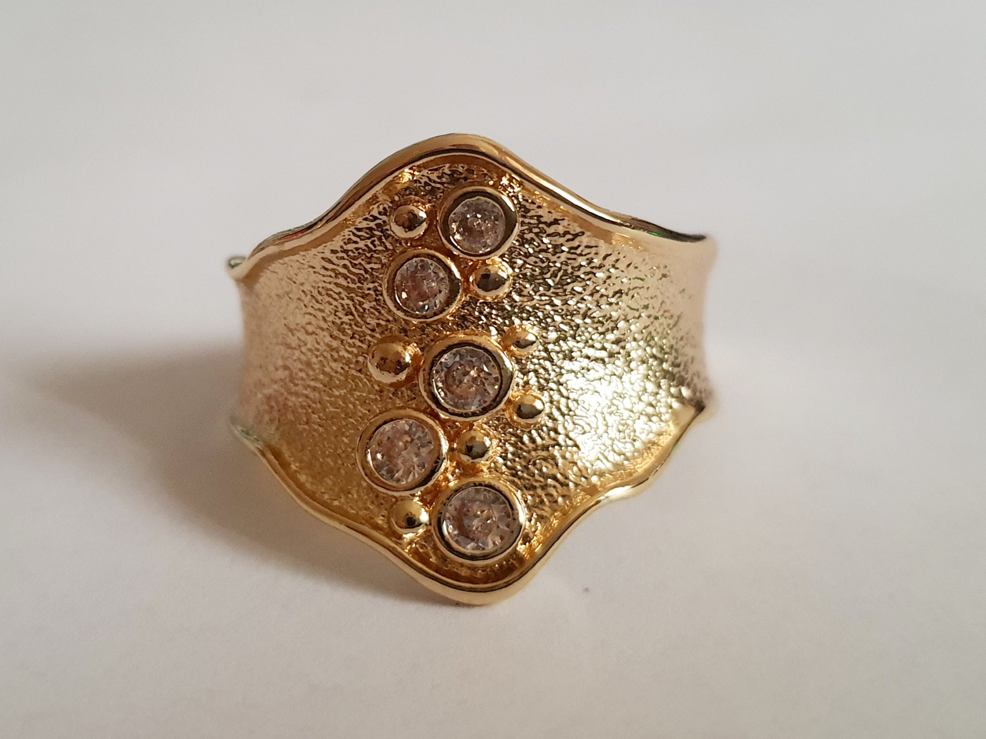 Szeroki złoty pierścionek ze stali nierdzewnej o młotkowanej fakturze, ozdobiony rzędem przezroczystych cyrkonii. Styl glamour, elegancja. Rozmiar 21 mm. Rouge Bizu.