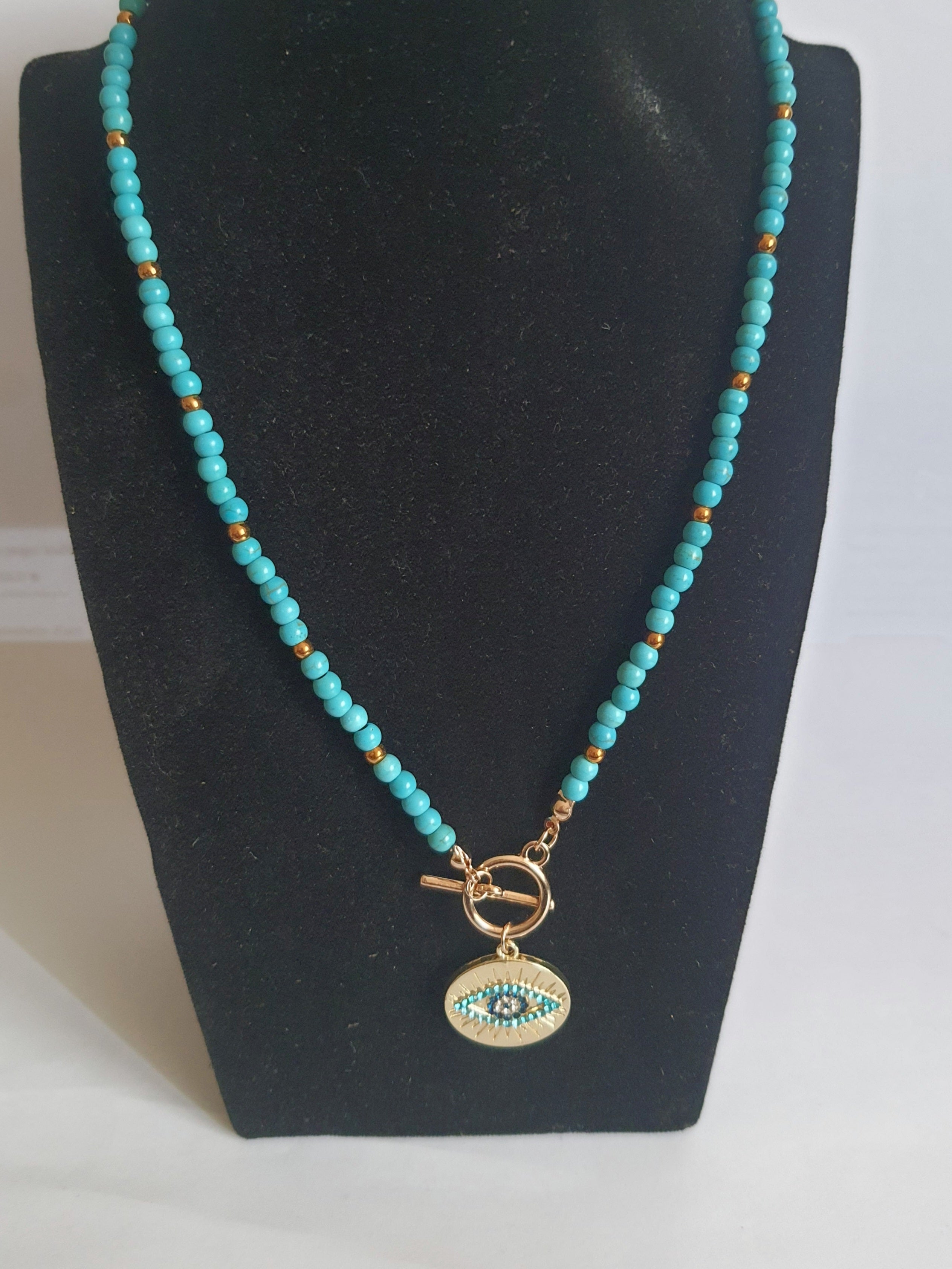 Naszyjnik choker z turkusowych koralików i złotych przekładek, z okrągłą złotą zawieszką z motywem oka w odcieniach turkusu i czerni. Długość 40 cm. Stal nierdzewna. Rouge Bizu.
