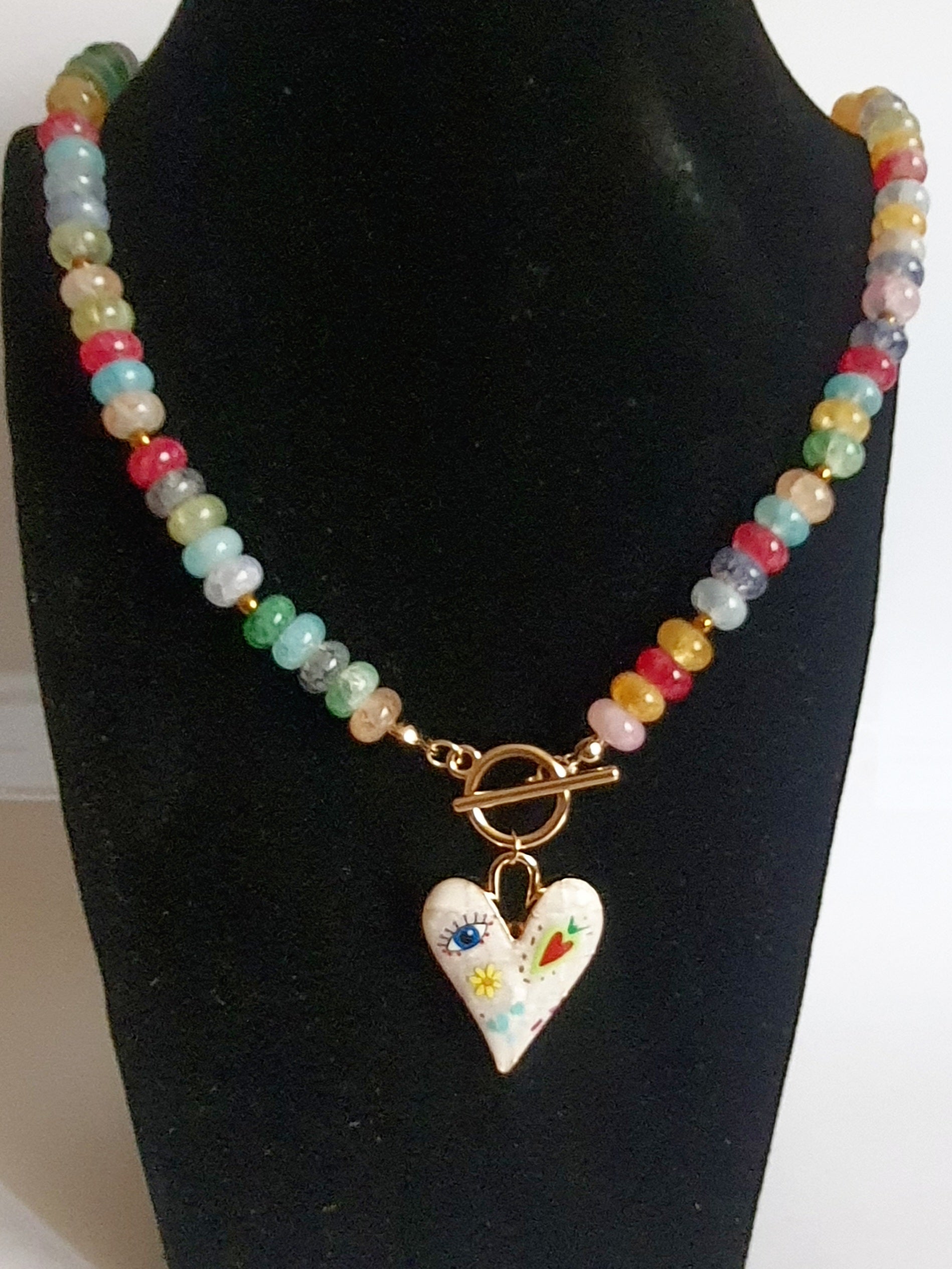 Naszyjnik z kolorowych koralików w stylu boho, z ozdobną zawieszką w kształcie białego serca zdobionego ręcznie malowanymi wzorami: oko, kwiat, serce i kolorowe symbole. Długość 45 cm. Stal nierdzewna. Rouge Bizu.