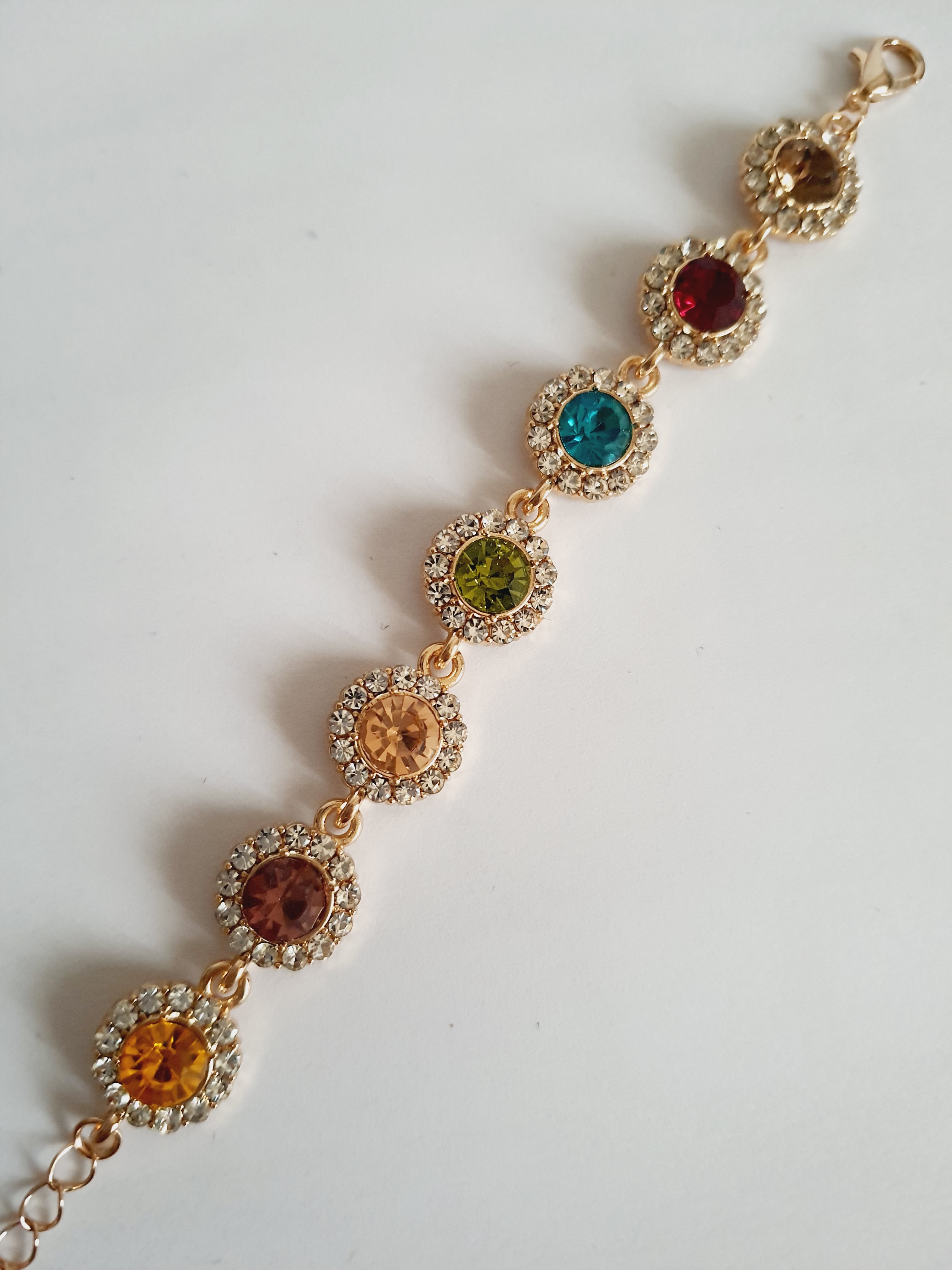 Bransoletka ze stali nierdzewnej w kolorze złota, z okrągłymi kolorowymi kryształami w odcieniach czerwieni, turkusu, zieleni, złota i bursztynu, otoczonymi małymi cyrkoniami. Długość regulowana 25 cm. Rouge Bizu.