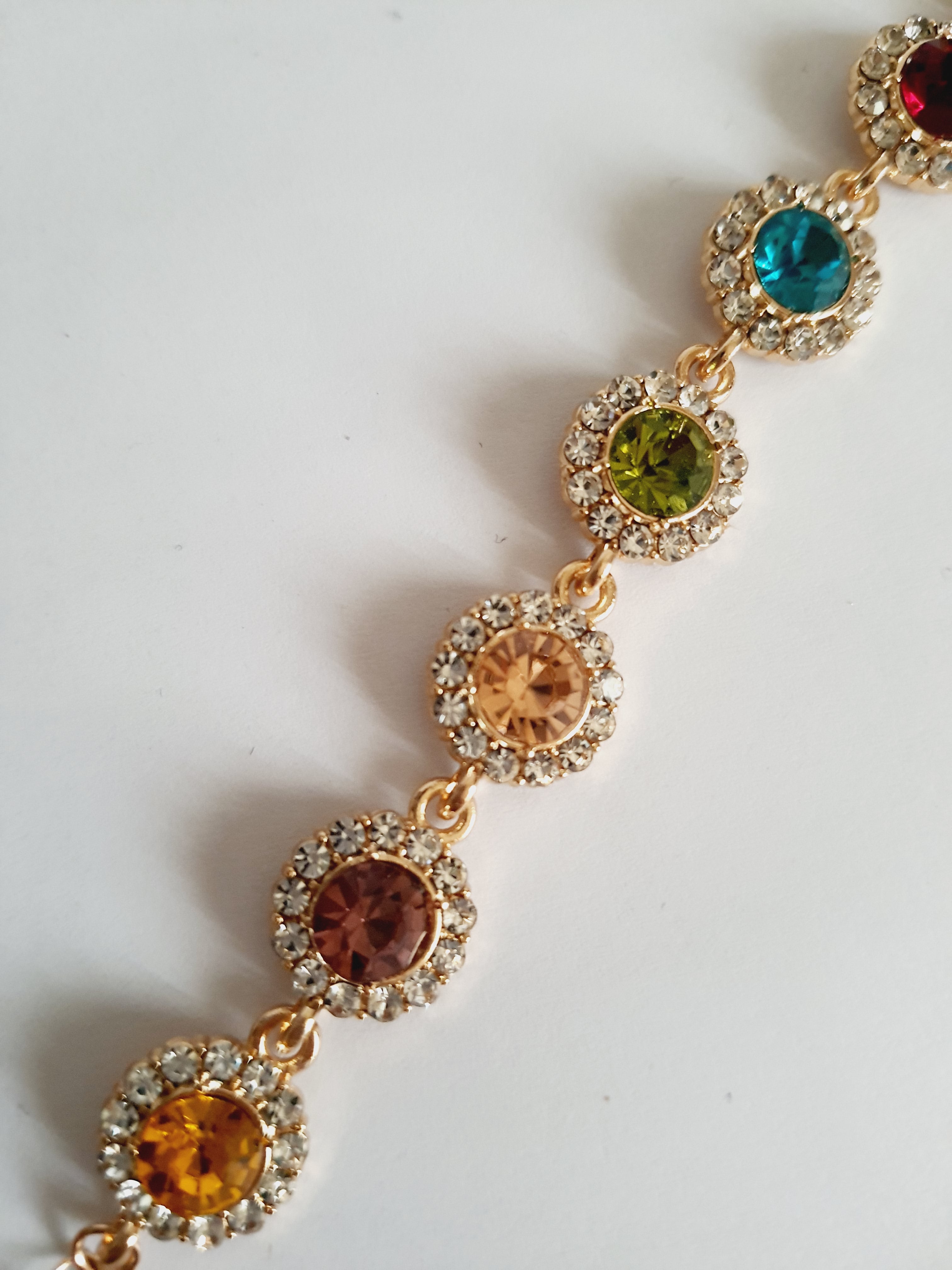 Bransoletka ze stali nierdzewnej w kolorze złota, z okrągłymi kolorowymi kryształami w odcieniach czerwieni, turkusu, zieleni, złota i bursztynu, otoczonymi małymi cyrkoniami. Długość regulowana 25 cm. Rouge Bizu.