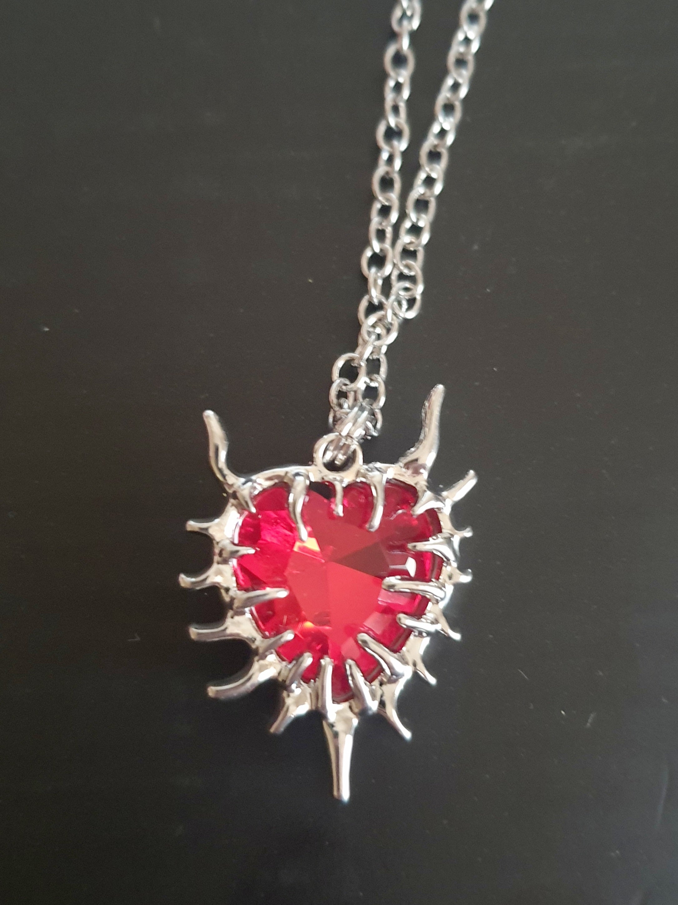 Naszyjnik z czerwonym kamieniem w kształcie serca, otoczonym srebrzystą oprawą przypominającą płomienie. Styl gotycki i wyrazisty. Długość łańcuszka 61 cm. Stal chirurgiczna, stal nierdzewna, wodoodporne, hipoalergiczne, jakość premium. RougeBizu.