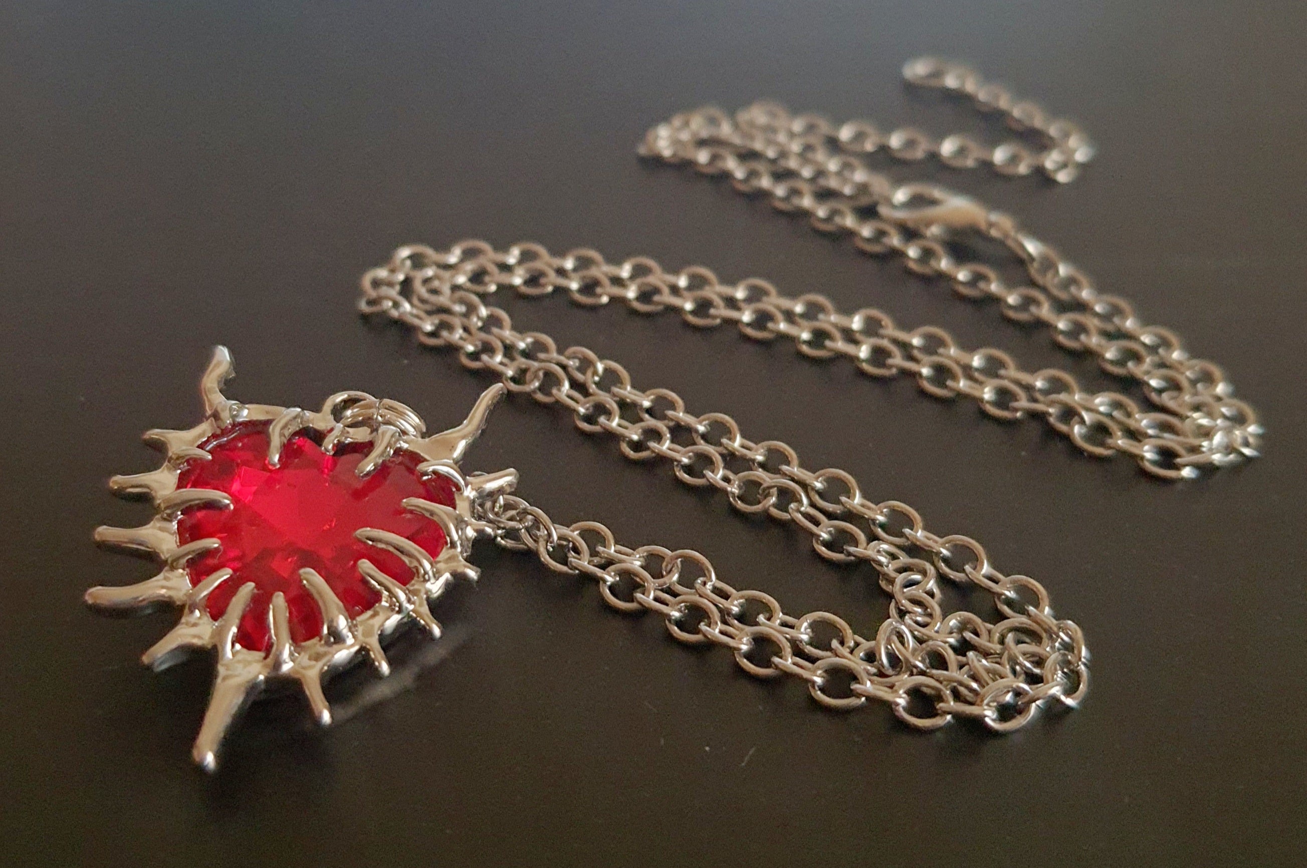 Naszyjnik z czerwonym kamieniem w kształcie serca, otoczonym srebrzystą oprawą przypominającą płomienie. Styl gotycki i wyrazisty. Długość łańcuszka 61 cm. Stal chirurgiczna, stal nierdzewna, wodoodporne, hipoalergiczne, jakość premium. RougeBizu.