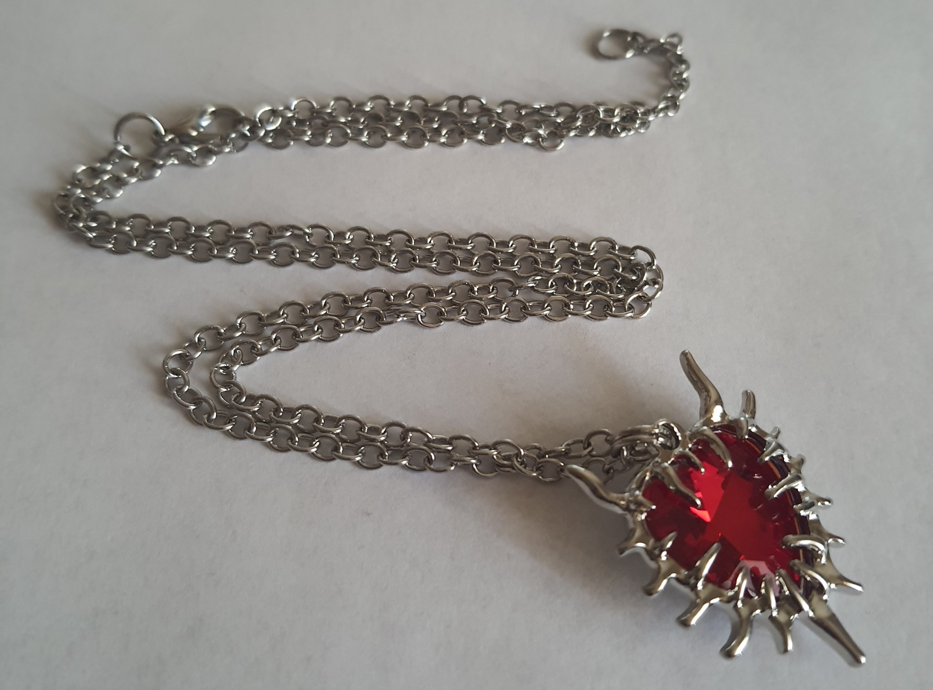 Naszyjnik z czerwonym kamieniem w kształcie serca, otoczonym srebrzystą oprawą przypominającą płomienie. Styl gotycki i wyrazisty. Długość łańcuszka 61 cm. Stal chirurgiczna, stal nierdzewna, wodoodporne, hipoalergiczne, jakość premium. RougeBizu.