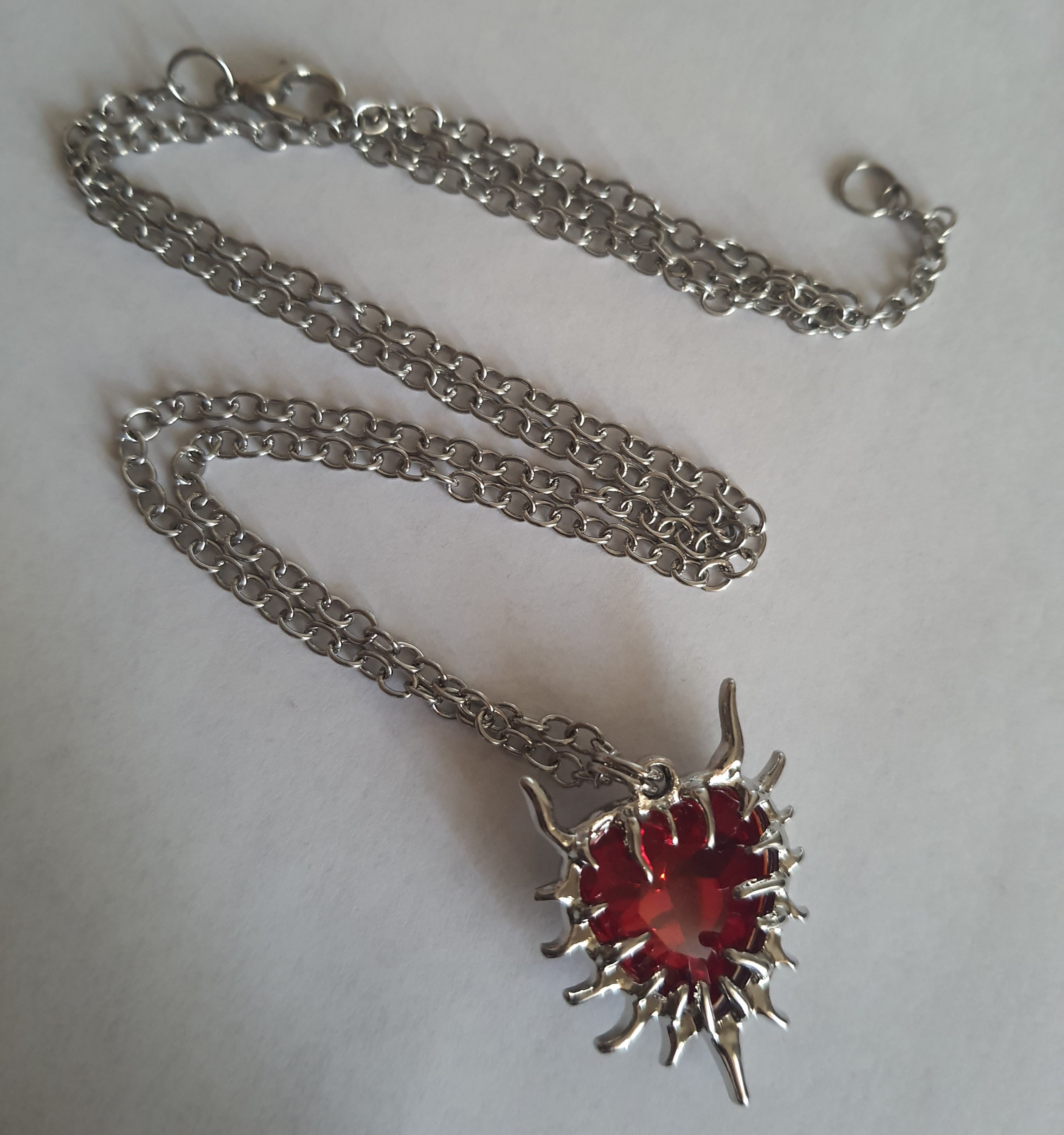 Naszyjnik z czerwonym kamieniem w kształcie serca, otoczonym srebrzystą oprawą przypominającą płomienie. Styl gotycki i wyrazisty. Długość łańcuszka 61 cm. Stal chirurgiczna, stal nierdzewna, wodoodporne, hipoalergiczne, jakość premium. RougeBizu.