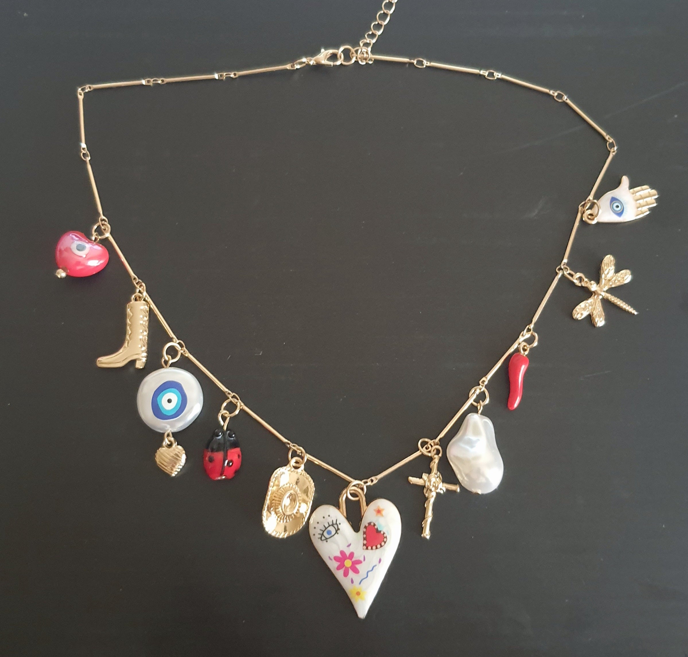 Złoty naszyjnik ze stali chirurgicznej z charmsami – sercem, biedronką, perłą syntetyczną, krzyżykiem, okiem proroczym, dłonią Fatimy, ważką i innymi symbolami. Regulowana długość 45–50 cm. Stal chirurgiczna, stal nierdzewna, wodoodporne, hipoalergiczne, jakość premium. RougeBizu.