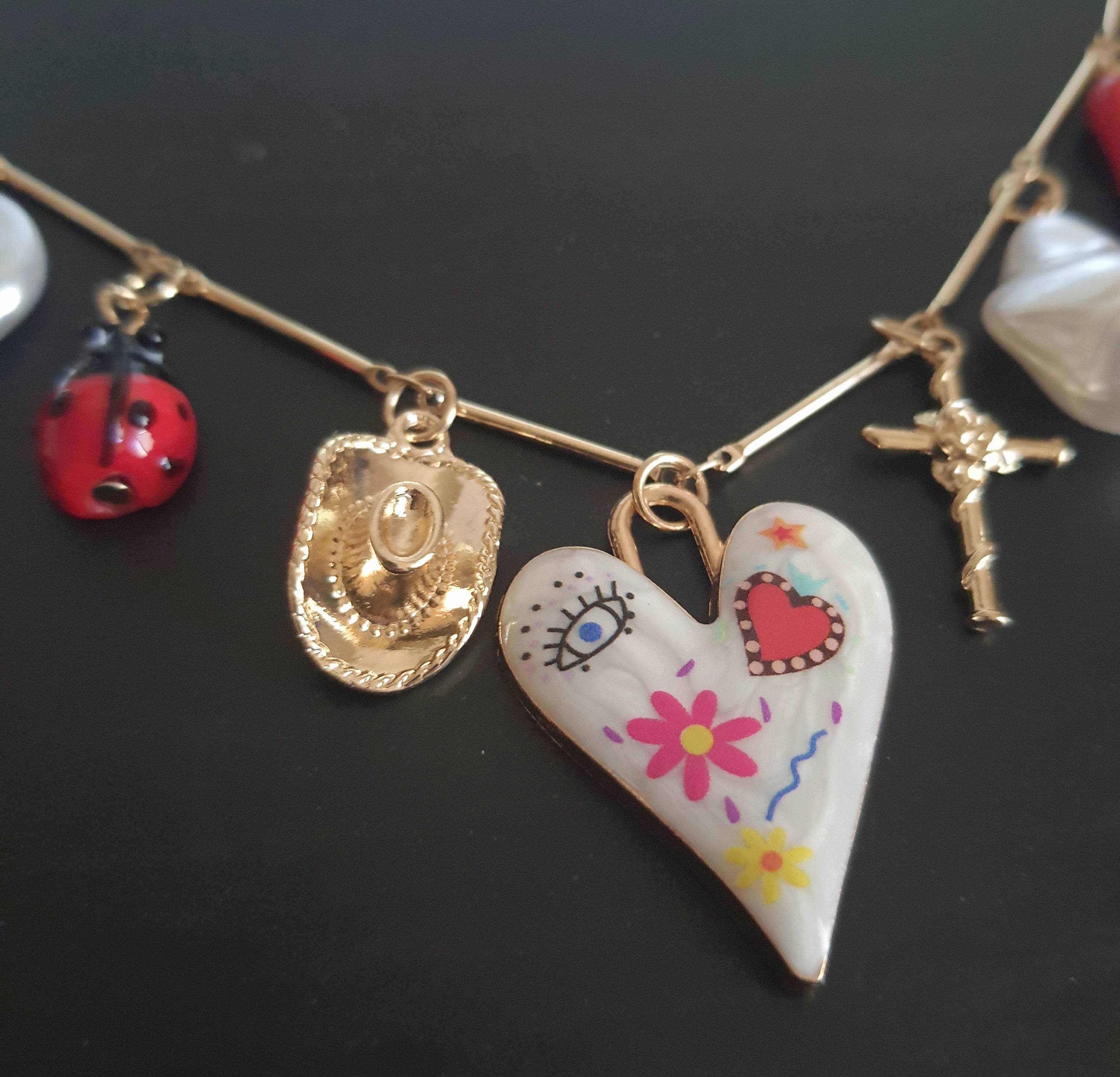 Złoty naszyjnik ze stali chirurgicznej z charmsami – sercem, biedronką, perłą syntetyczną, krzyżykiem, okiem proroczym, dłonią Fatimy, ważką i innymi symbolami. Regulowana długość 45–50 cm. Stal chirurgiczna, stal nierdzewna, wodoodporne, hipoalergiczne, jakość premium. RougeBizu.