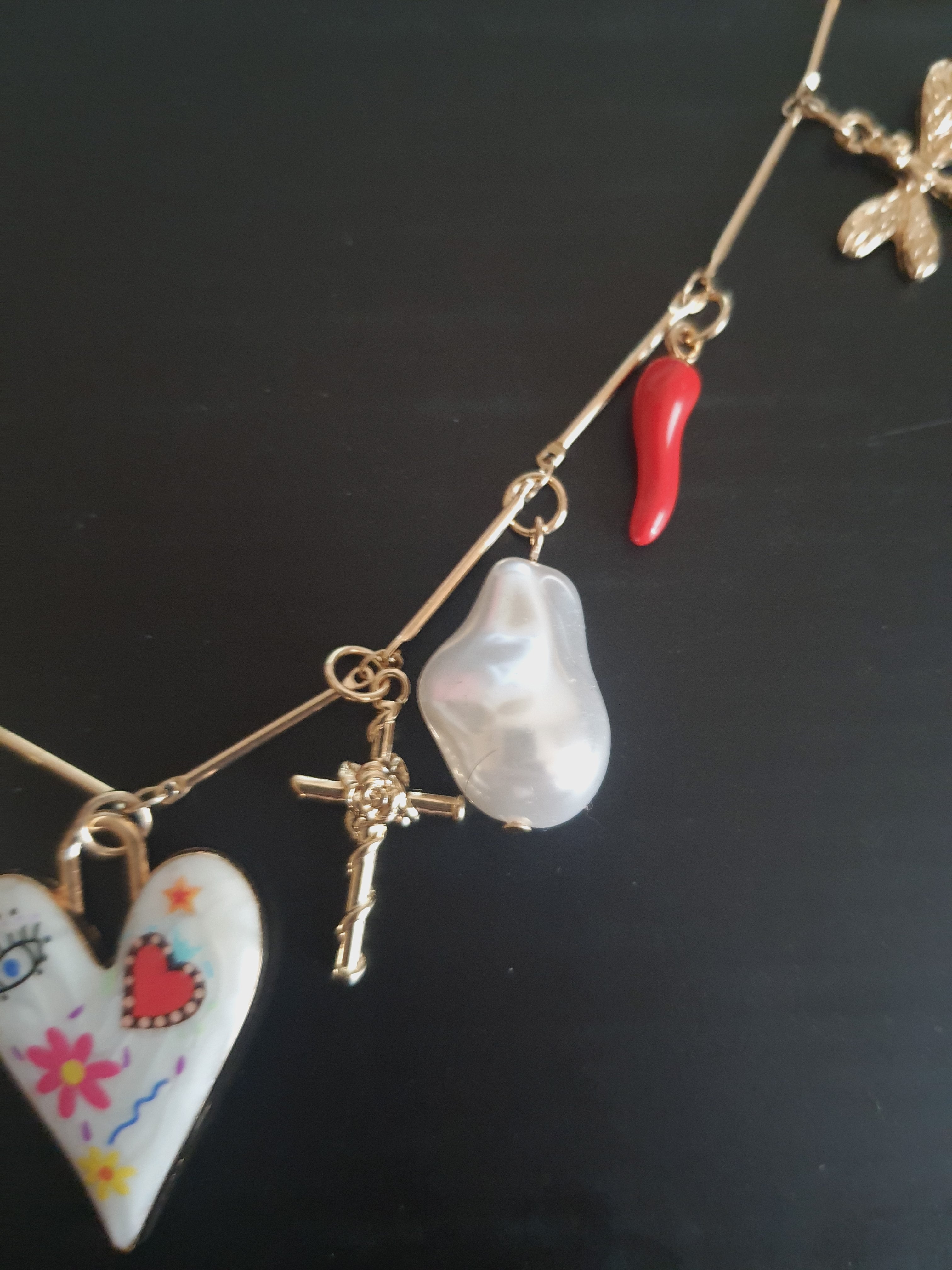 Złoty naszyjnik ze stali chirurgicznej z charmsami – sercem, biedronką, perłą syntetyczną, krzyżykiem, okiem proroczym, dłonią Fatimy, ważką i innymi symbolami. Regulowana długość 45–50 cm. Stal chirurgiczna, stal nierdzewna, wodoodporne, hipoalergiczne, jakość premium. RougeBizu.