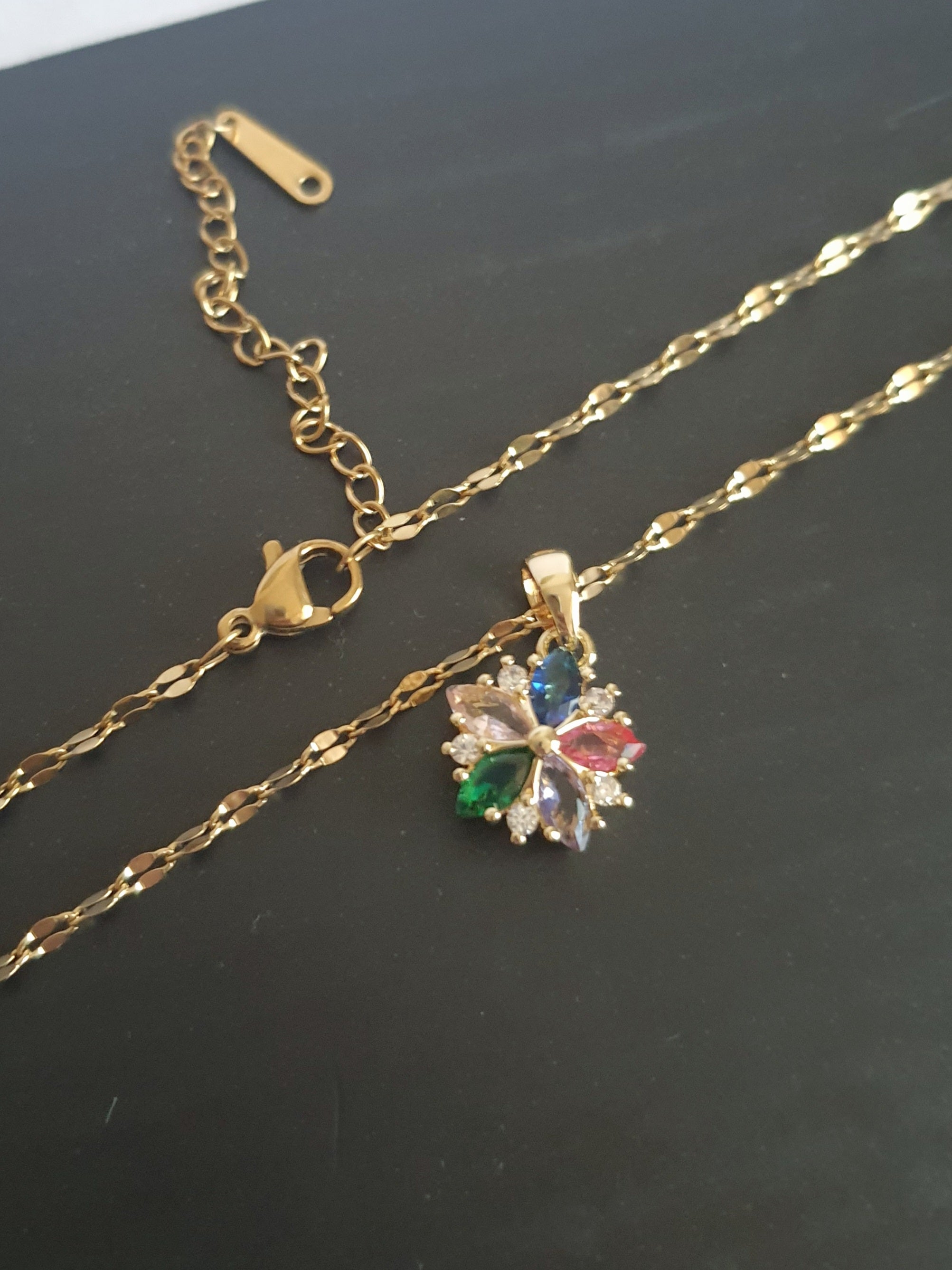 Złoty naszyjnik ze stali chirurgicznej z zawieszką w kształcie kwiatka wysadzanego wielokolorowymi cyrkoniami (zielone, różowe, błękitne, granatowe). Regulowana długość 40–45 cm. Stal chirurgiczna, stal nierdzewna, wodoodporne, hipoalergiczne, jakość premium. RougeBizu.