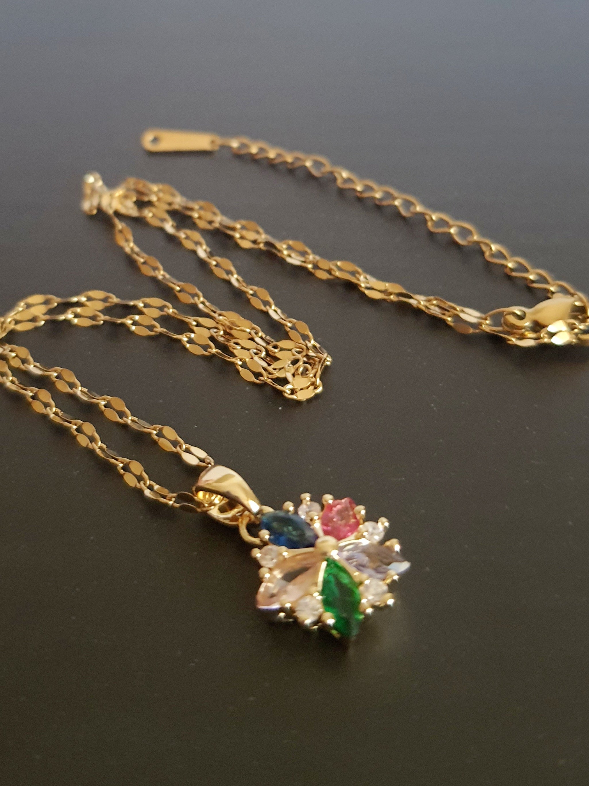 Złoty naszyjnik ze stali chirurgicznej z zawieszką w kształcie kwiatka wysadzanego wielokolorowymi cyrkoniami (zielone, różowe, błękitne, granatowe). Regulowana długość 40–45 cm. Stal chirurgiczna, stal nierdzewna, wodoodporne, hipoalergiczne, jakość premium. RougeBizu.