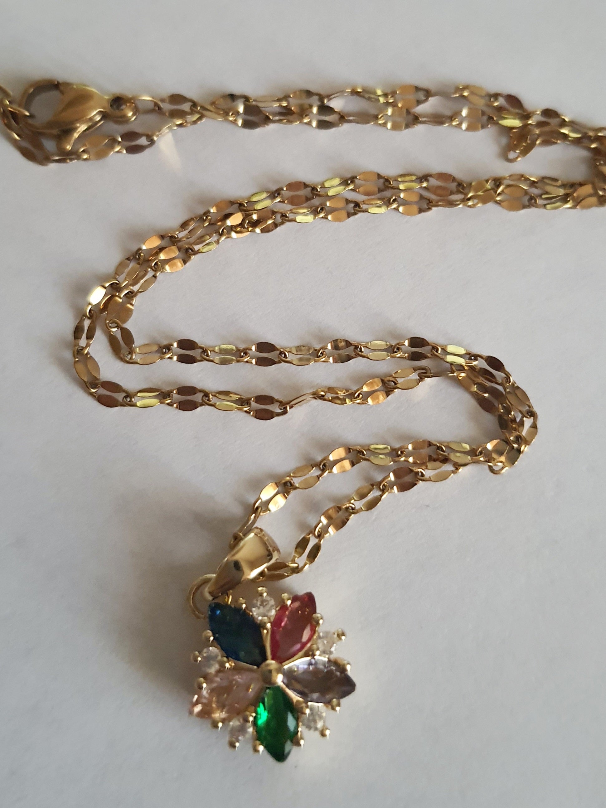 Złoty naszyjnik ze stali chirurgicznej z zawieszką w kształcie kwiatka wysadzanego wielokolorowymi cyrkoniami (zielone, różowe, błękitne, granatowe). Regulowana długość 40–45 cm. Stal chirurgiczna, stal nierdzewna, wodoodporne, hipoalergiczne, jakość premium. RougeBizu.