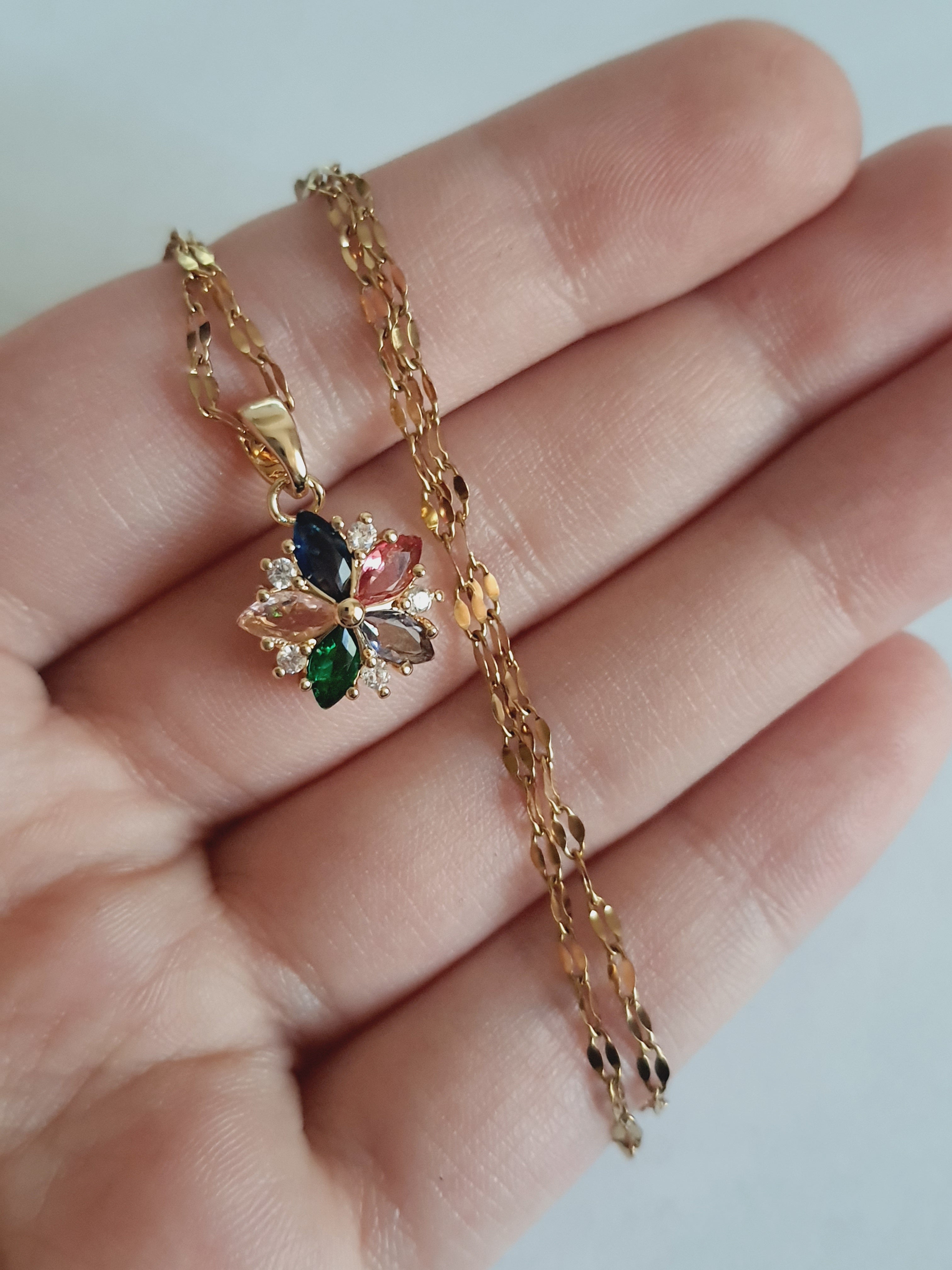 Złoty naszyjnik ze stali chirurgicznej z zawieszką w kształcie kwiatka wysadzanego wielokolorowymi cyrkoniami (zielone, różowe, błękitne, granatowe). Regulowana długość 40–45 cm. Stal chirurgiczna, stal nierdzewna, wodoodporne, hipoalergiczne, jakość premium. RougeBizu.