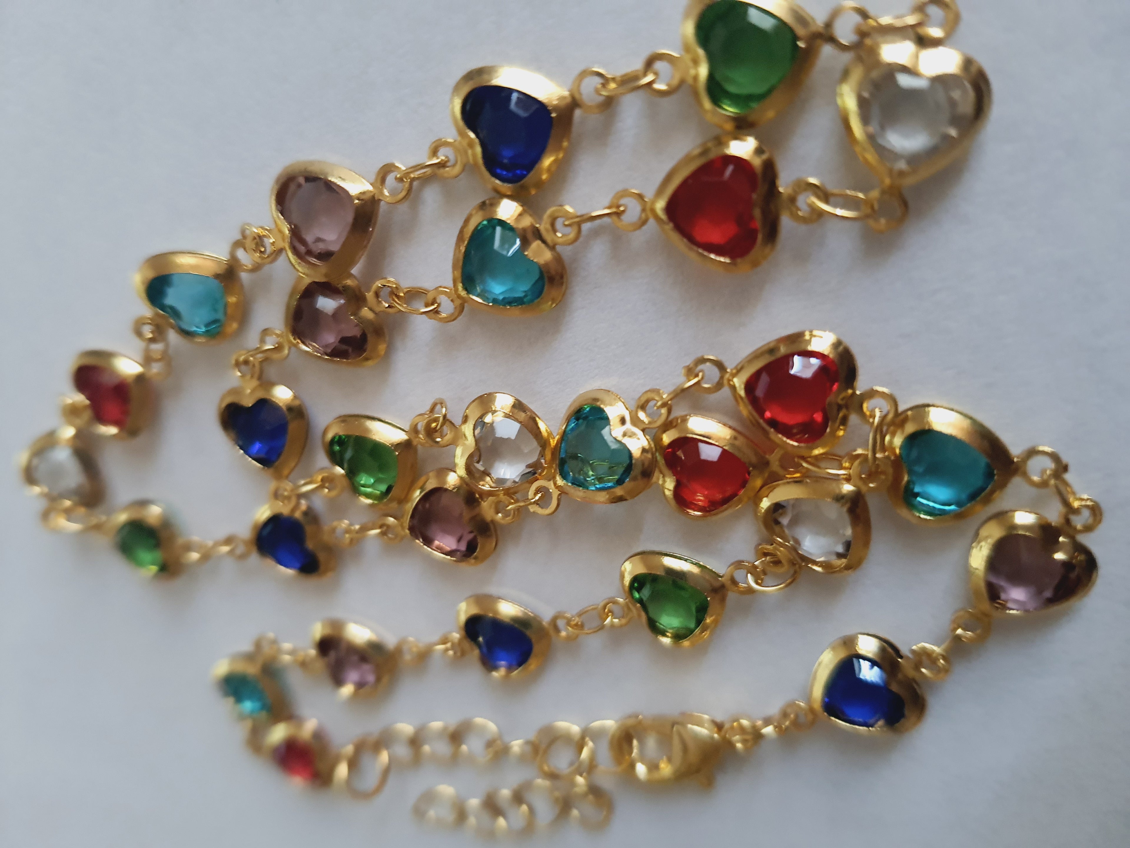 Złoty naszyjnik ze stali chirurgicznej z kolorowymi serduszkami z kryształów w barwach czerwonych, zielonych, niebieskich, fioletowych i turkusowych. Regulowana długość 40–45 cm. Stal chirurgiczna, stal nierdzewna, wodoodporne, hipoalergiczne, jakość premium. RougeBizu.