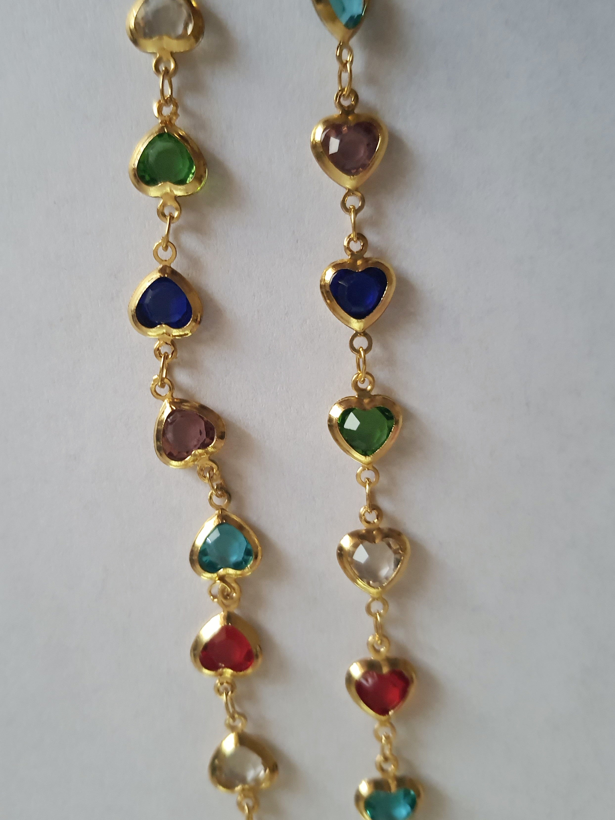 Złoty naszyjnik ze stali chirurgicznej z kolorowymi serduszkami z kryształów w barwach czerwonych, zielonych, niebieskich, fioletowych i turkusowych. Regulowana długość 40–45 cm. Stal chirurgiczna, stal nierdzewna, wodoodporne, hipoalergiczne, jakość premium. RougeBizu.