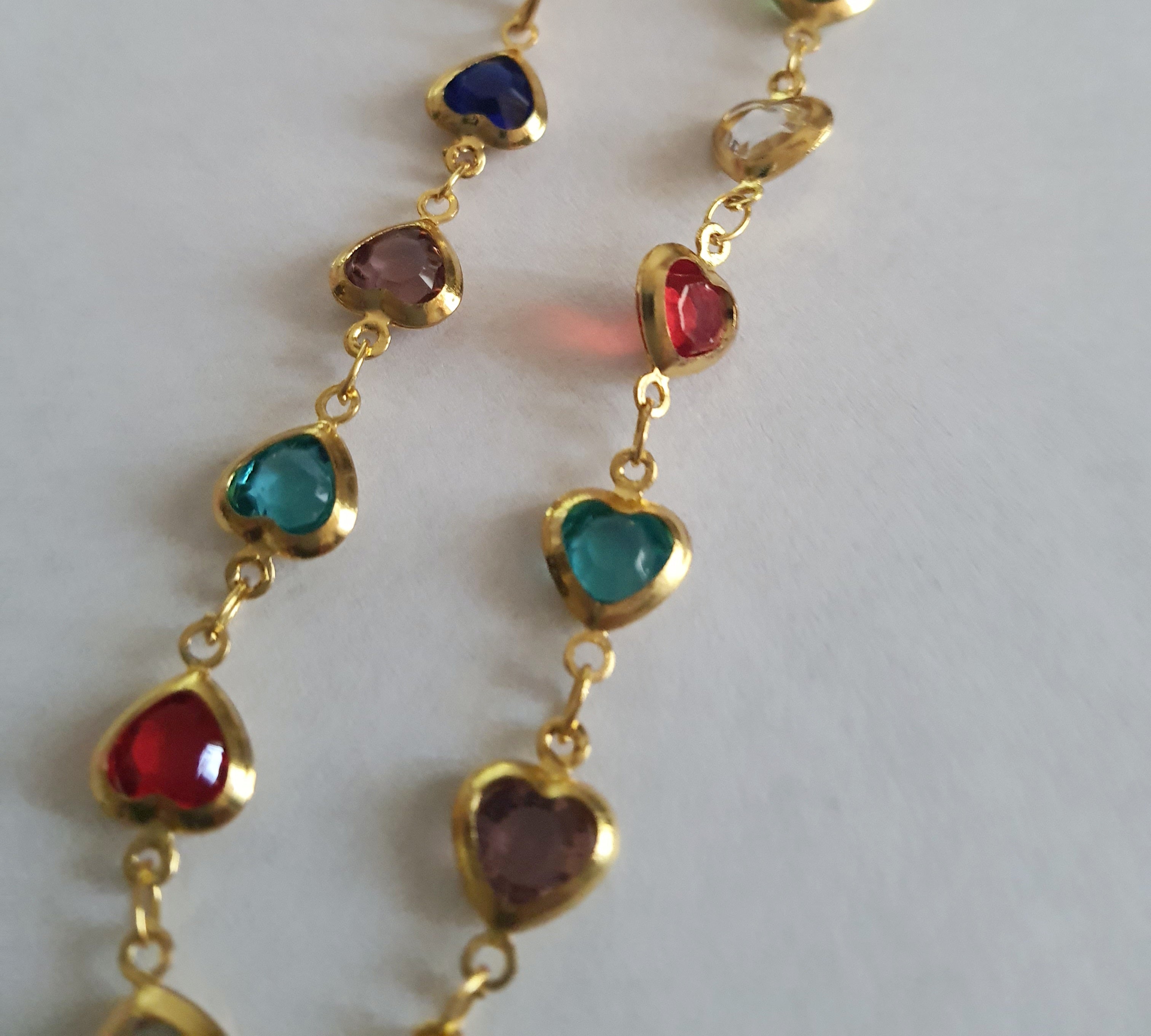 Złoty naszyjnik ze stali chirurgicznej z kolorowymi serduszkami z kryształów w barwach czerwonych, zielonych, niebieskich, fioletowych i turkusowych. Regulowana długość 40–45 cm. Stal chirurgiczna, stal nierdzewna, wodoodporne, hipoalergiczne, jakość premium. RougeBizu.