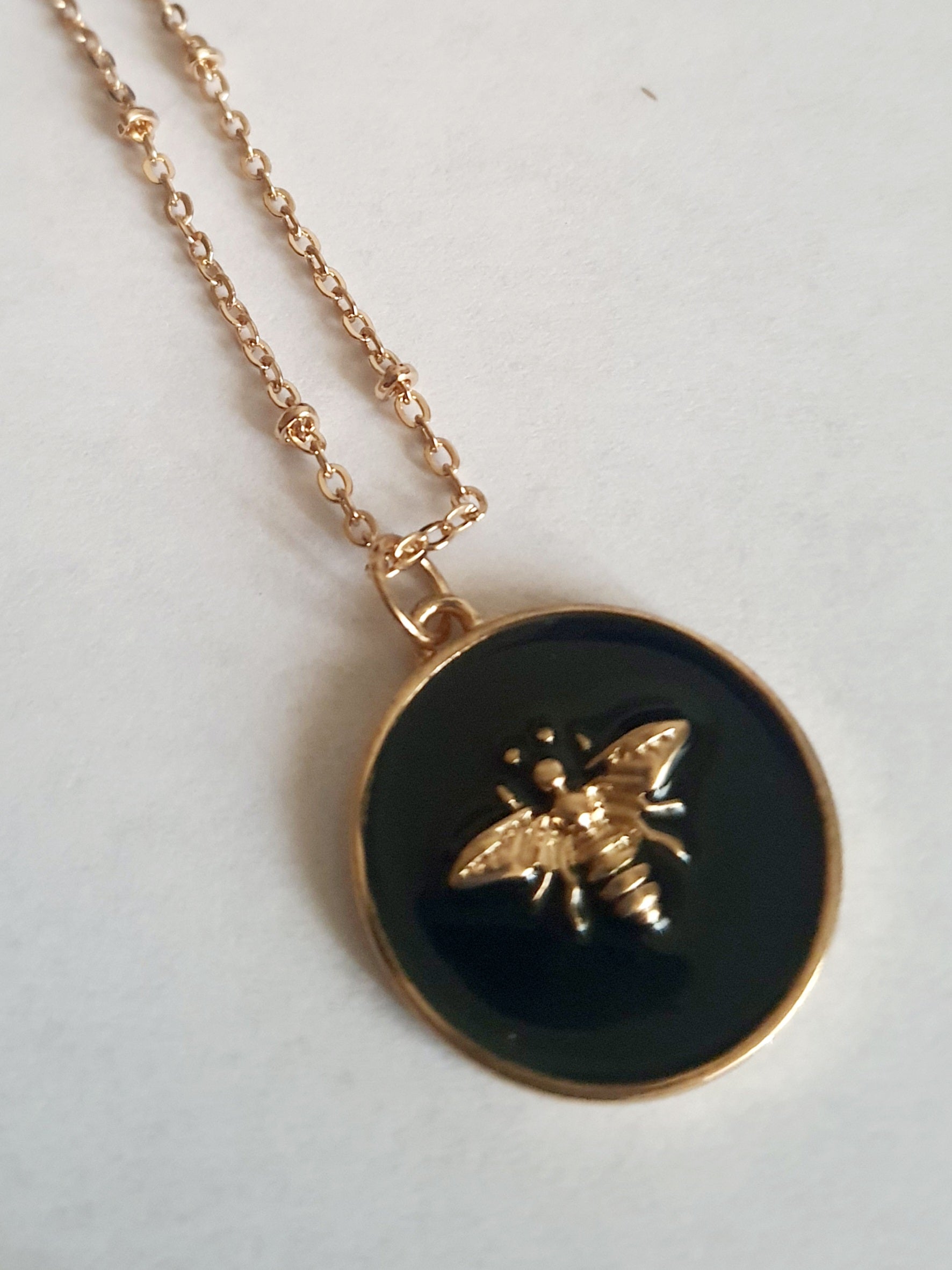 Złoty naszyjnik ze stali chirurgicznej z czarną okrągłą zawieszką i złotą pszczółką. Regulowana długość 40–49 cm. Stal chirurgiczna, stal nierdzewna, wodoodporne, hipoalergiczne, jakość premium. RougeBizu.