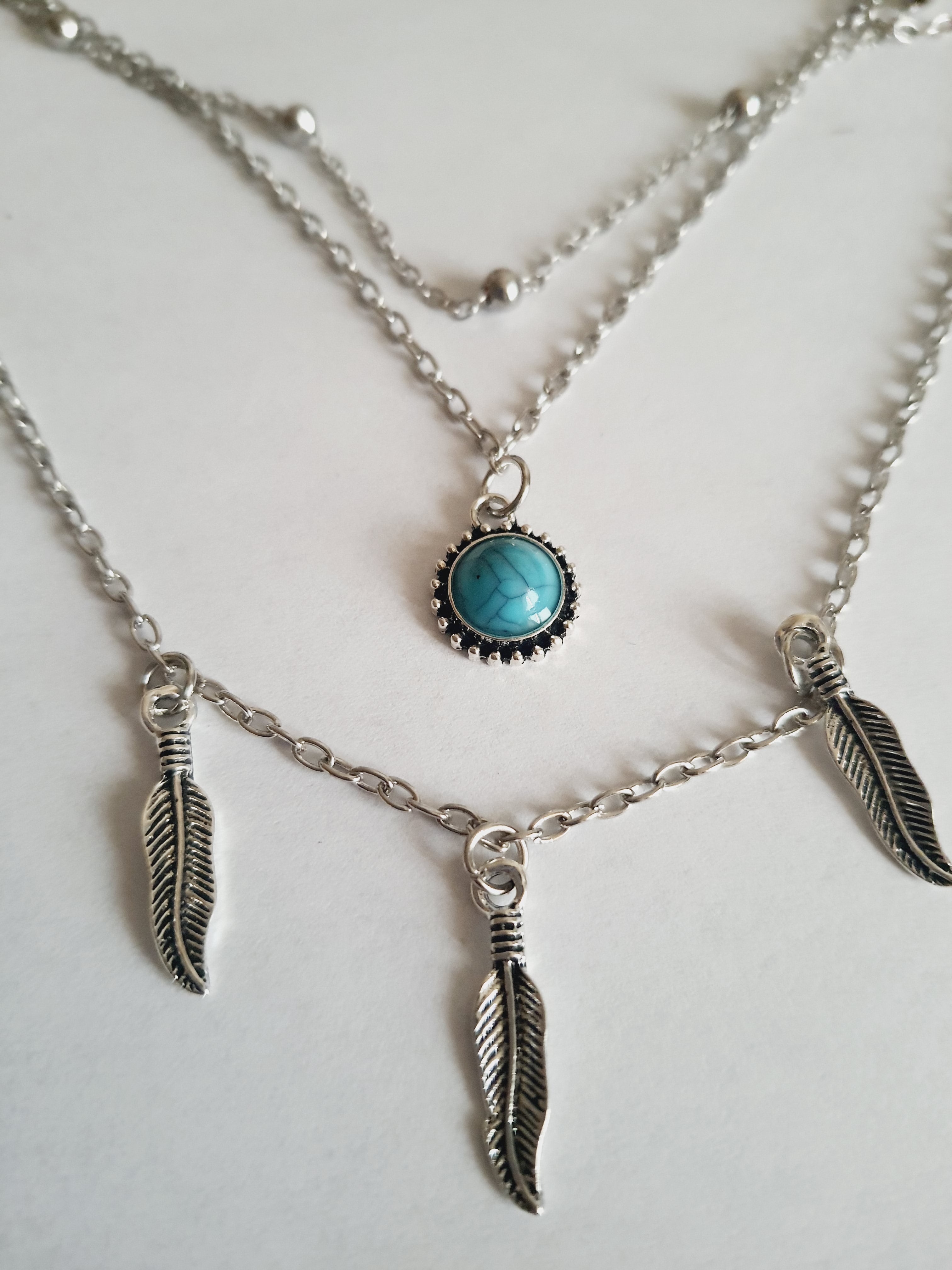 Srebrny potrójny naszyjnik ze stali chirurgicznej – jeden łańcuszek z piórkami, drugi z turkusowym kamieniem, trzeci z kuleczkami. Regulowana długość 39–45 cm. Stal chirurgiczna, stal nierdzewna, wodoodporne, hipoalergiczne, jakość premium. RougeBizu.