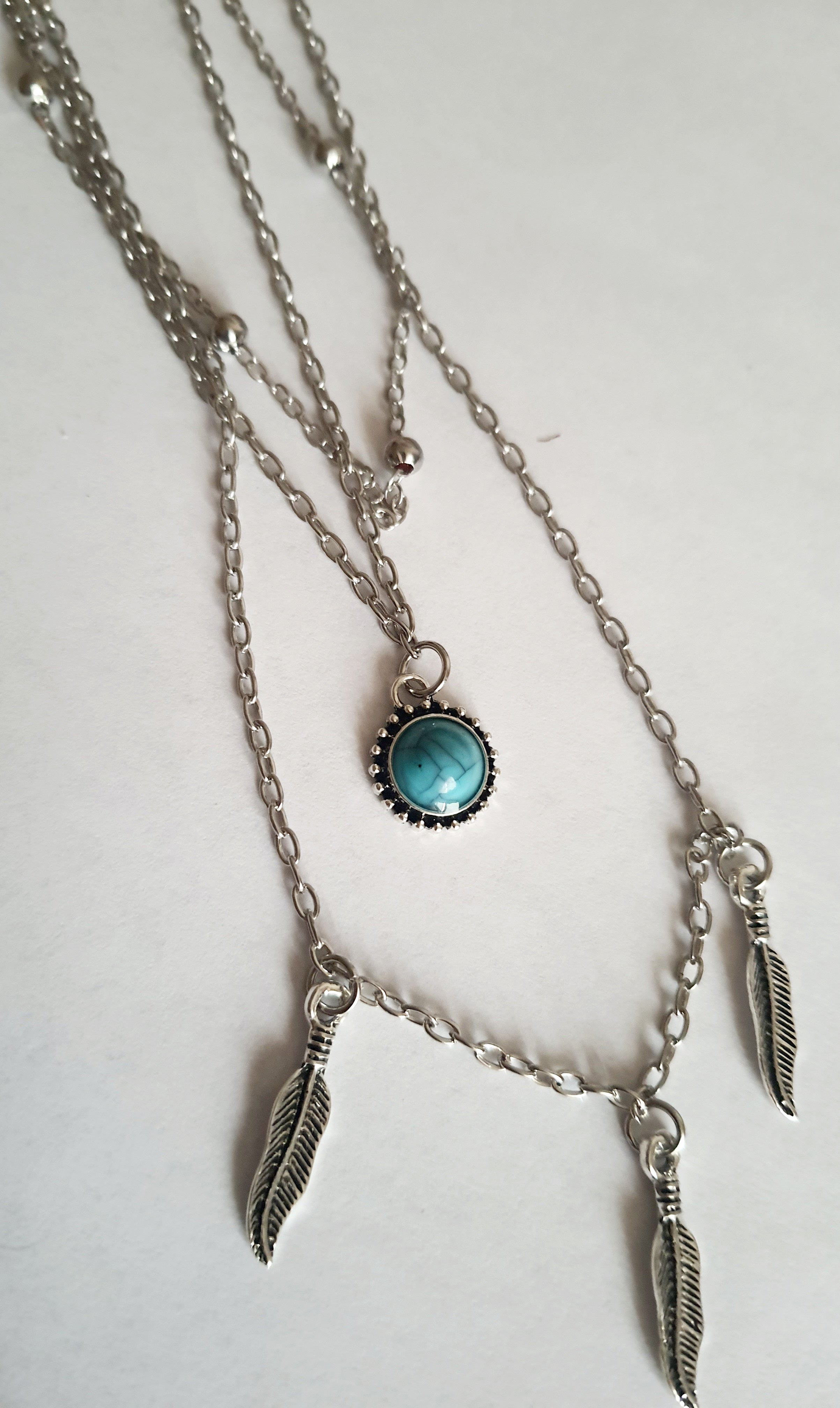 Srebrny potrójny naszyjnik ze stali chirurgicznej – jeden łańcuszek z piórkami, drugi z turkusowym kamieniem, trzeci z kuleczkami. Regulowana długość 39–45 cm. Stal chirurgiczna, stal nierdzewna, wodoodporne, hipoalergiczne, jakość premium. RougeBizu.