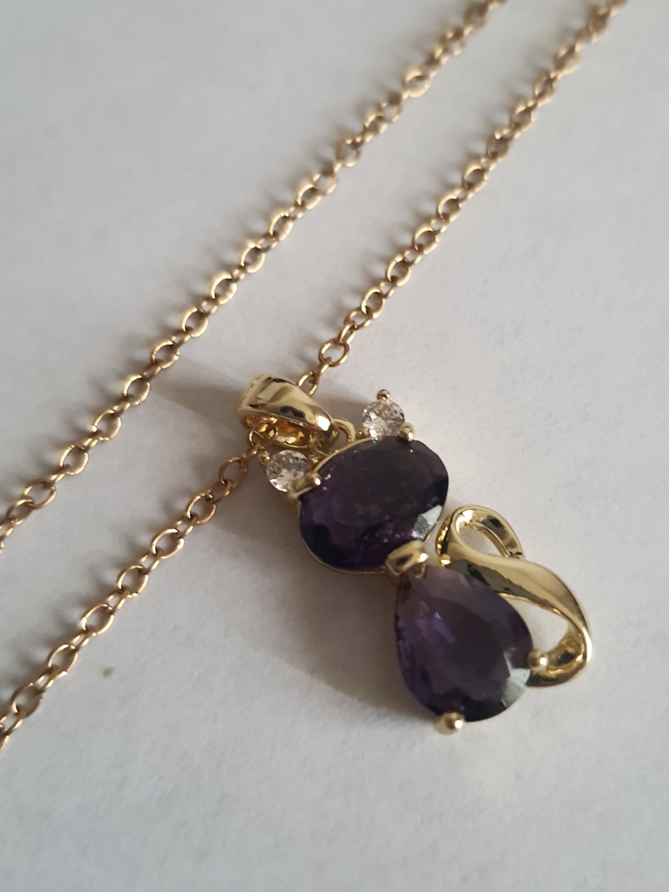 Naszyjnik złoty ze stali nierdzewnej – stal chirurgiczna, wodoodporny, hipoalergiczny, jakość premium. Zawieszka w kształcie kotka z dwóch fioletowych kryształów, ozdobiona złotym detalem i drobnymi cyrkoniami. Długość łańcuszka 40–45 cm. Elegancja i styl RougeBizu.