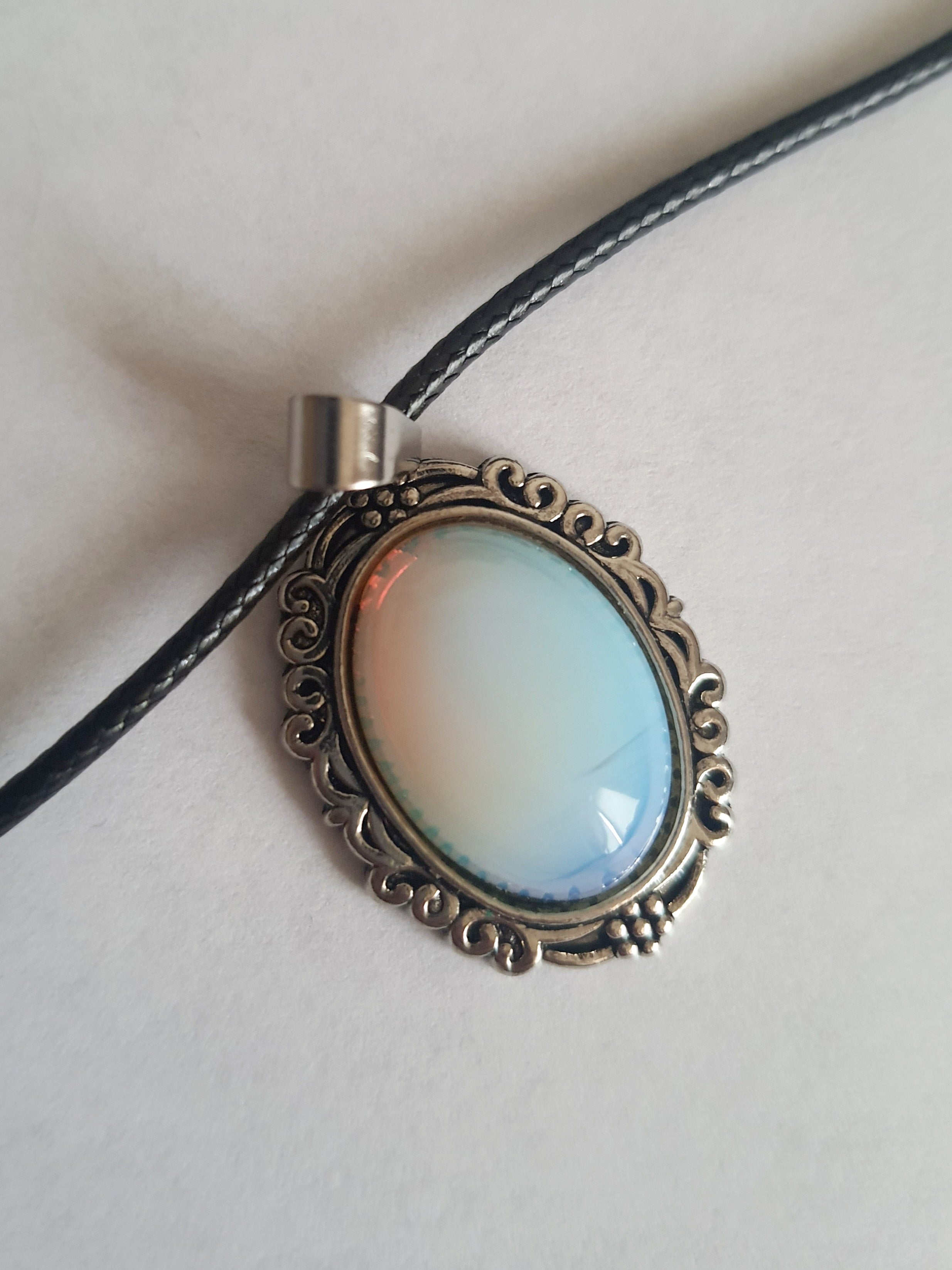 Naszyjnik vintage ze stali nierdzewnej – stal chirurgiczna, wodoodporny, hipoalergiczny, jakość premium. Zawieszka owalna z syntetycznym opalem opalizującym błękitem i bielą, w zdobionej ramie. Czarna linka o długości 45–50 cm. Styl RougeBizu.