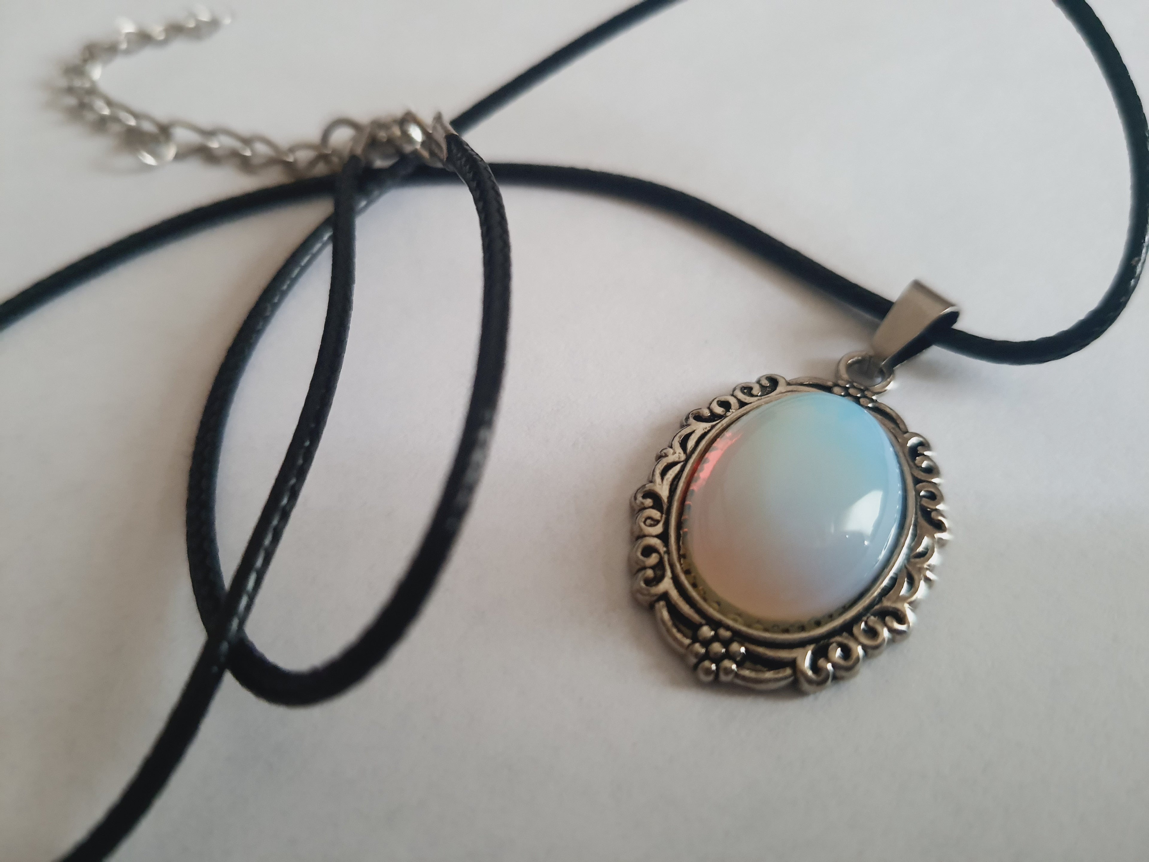 Naszyjnik vintage ze stali nierdzewnej – stal chirurgiczna, wodoodporny, hipoalergiczny, jakość premium. Zawieszka owalna z syntetycznym opalem opalizującym błękitem i bielą, w zdobionej ramie. Czarna linka o długości 45–50 cm. Styl RougeBizu.