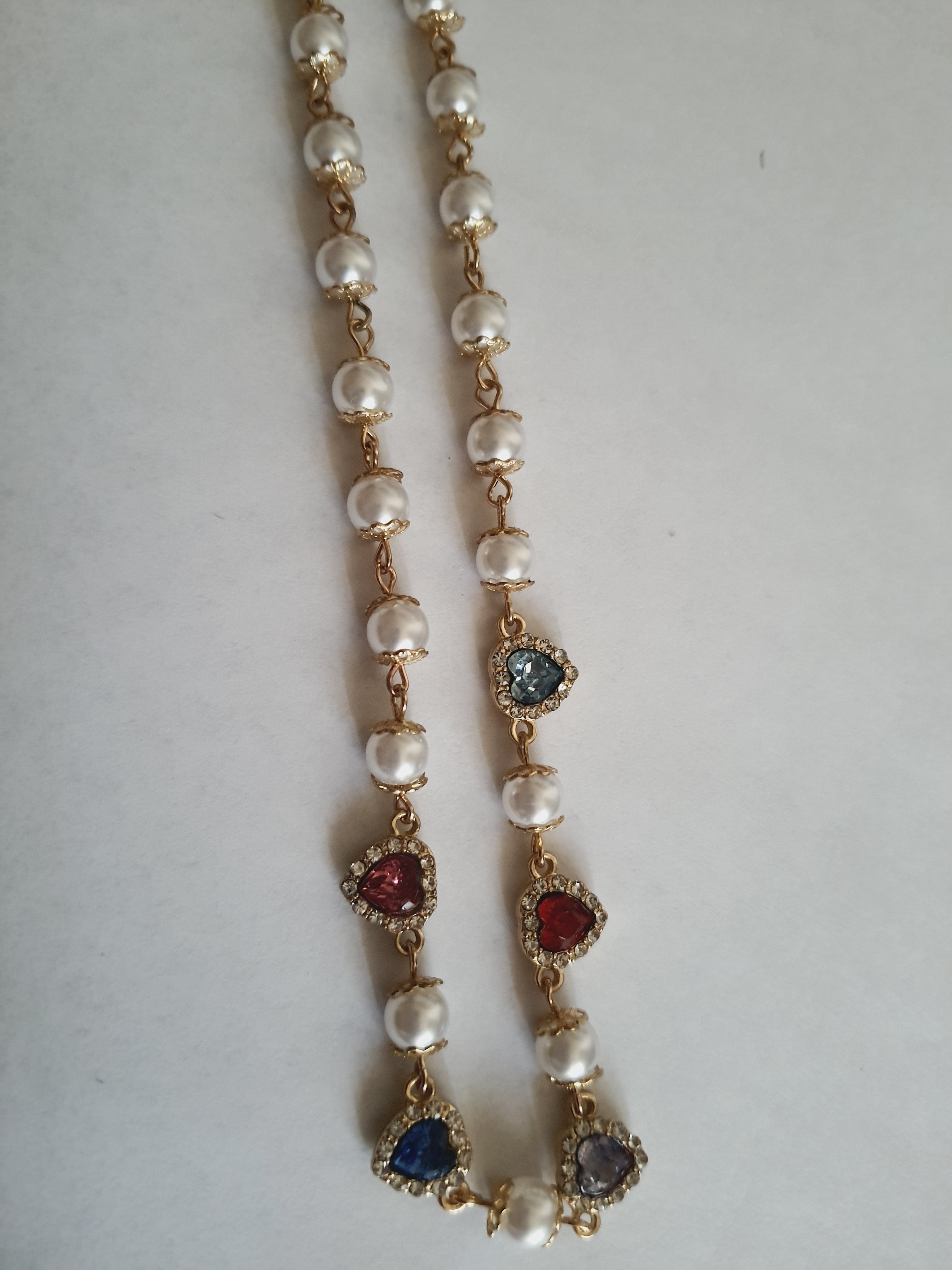 Naszyjnik złoty ze stali nierdzewnej – stal chirurgiczna, wodoodporny, hipoalergiczny, jakość premium. Perły syntetyczne i zawieszki w kształcie kolorowych serc (czerwony, niebieski, różowy, fioletowy) zdobione cyrkoniami. Długość 40–49 cm. RougeBizu.