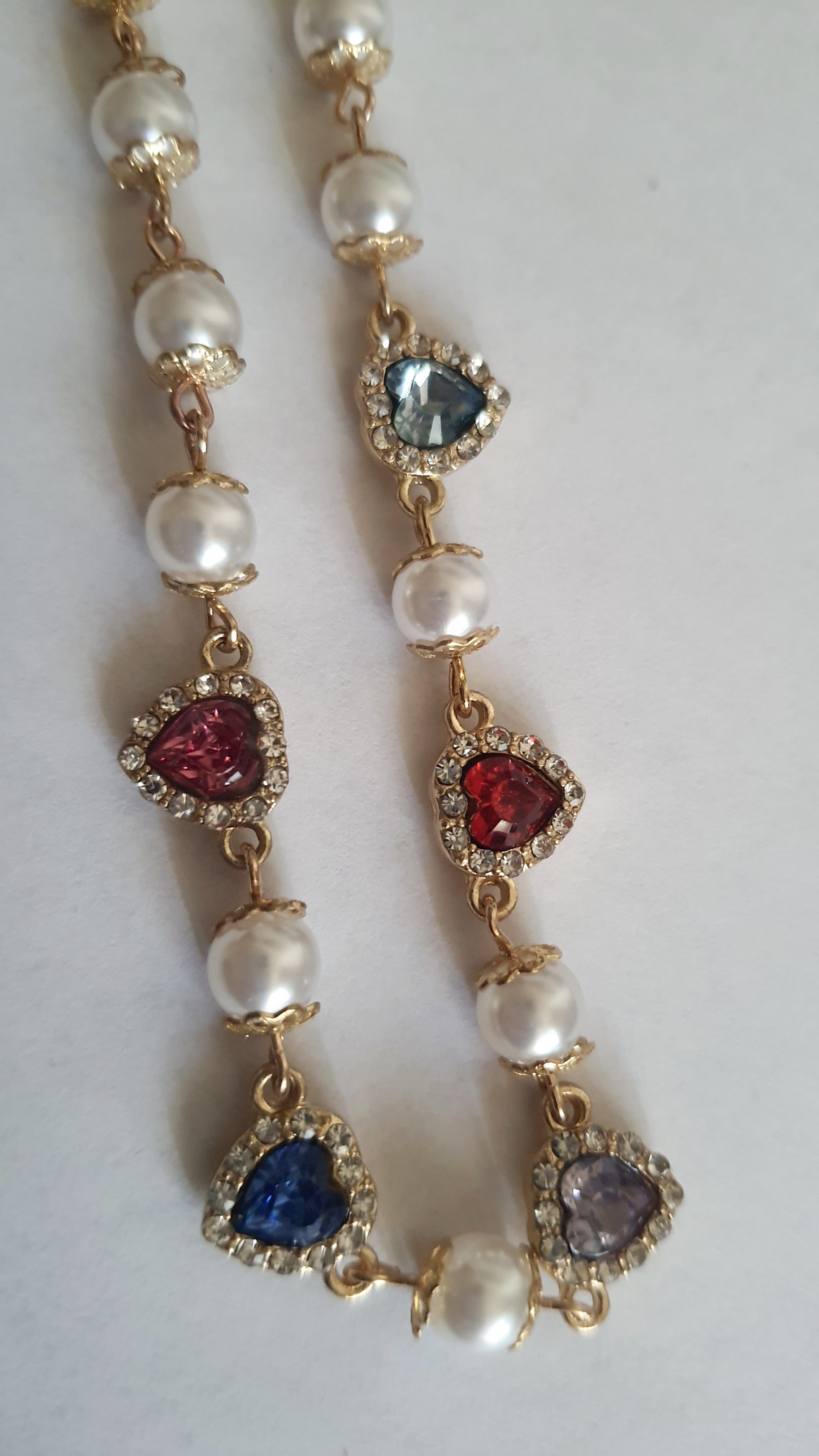 Naszyjnik złoty ze stali nierdzewnej – stal chirurgiczna, wodoodporny, hipoalergiczny, jakość premium. Perły syntetyczne i zawieszki w kształcie kolorowych serc (czerwony, niebieski, różowy, fioletowy) zdobione cyrkoniami. Długość 40–49 cm. RougeBizu.