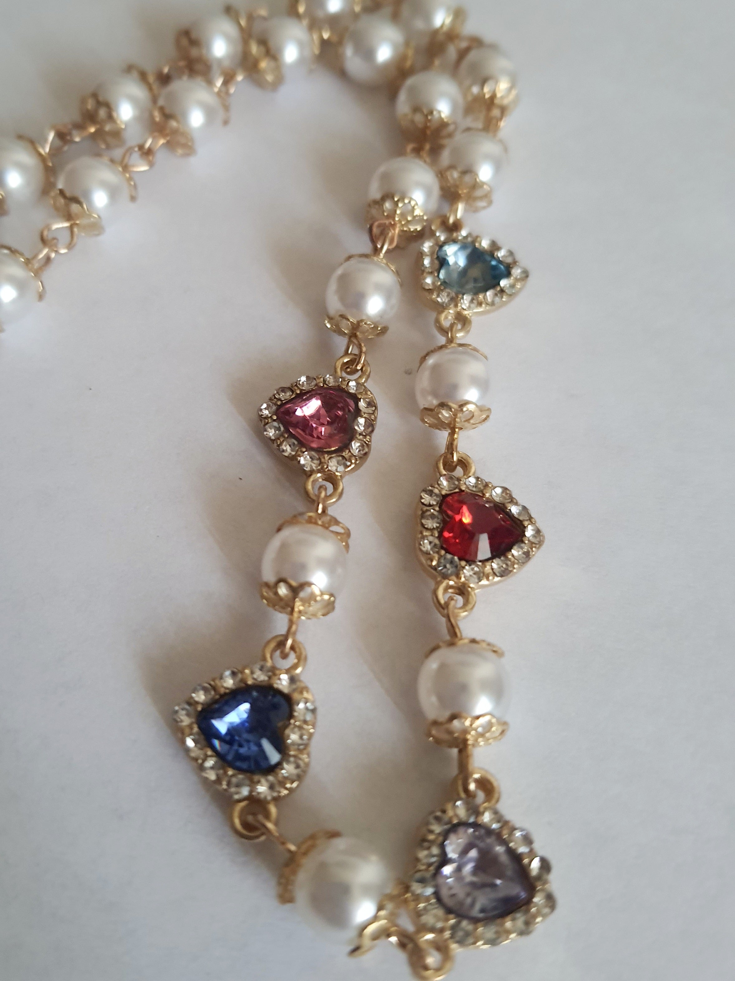 Naszyjnik złoty ze stali nierdzewnej – stal chirurgiczna, wodoodporny, hipoalergiczny, jakość premium. Perły syntetyczne i zawieszki w kształcie kolorowych serc (czerwony, niebieski, różowy, fioletowy) zdobione cyrkoniami. Długość 40–49 cm. RougeBizu.