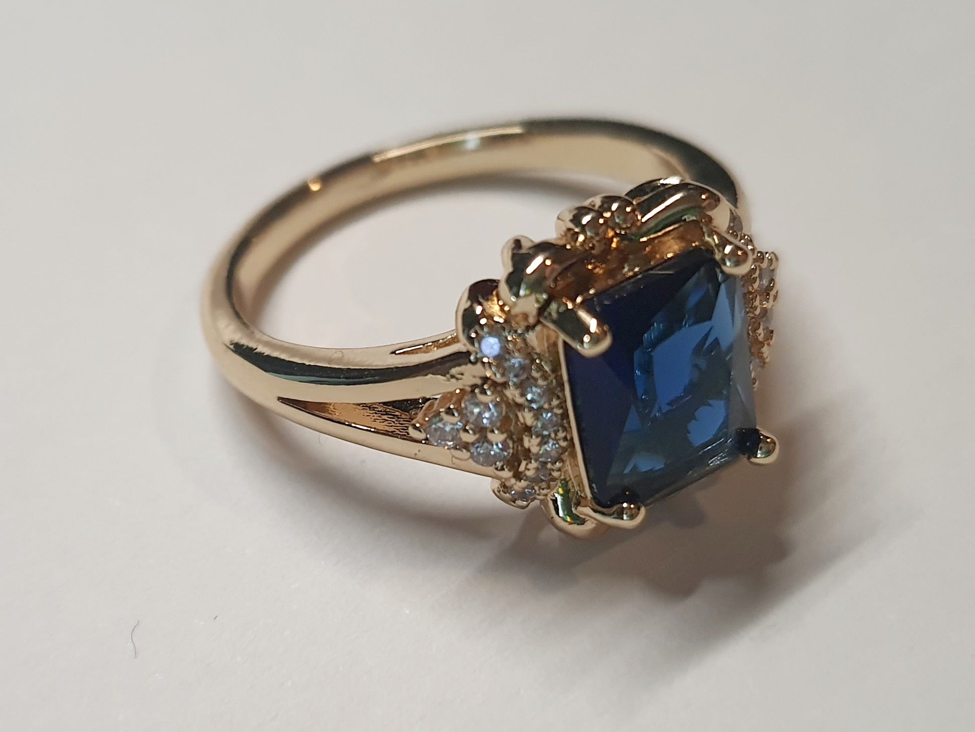 Złoty pierścionek ze stali nierdzewnej z granatowym kamieniem w szlifie szmaragdowym i cyrkoniami po bokach w stylu glamour klasycznym. Rozmiar 20 mm. stal chirurgiczna, stal nierdzewna, wodoodporne, hipoalergiczne, jakość premium, RougeBizu.