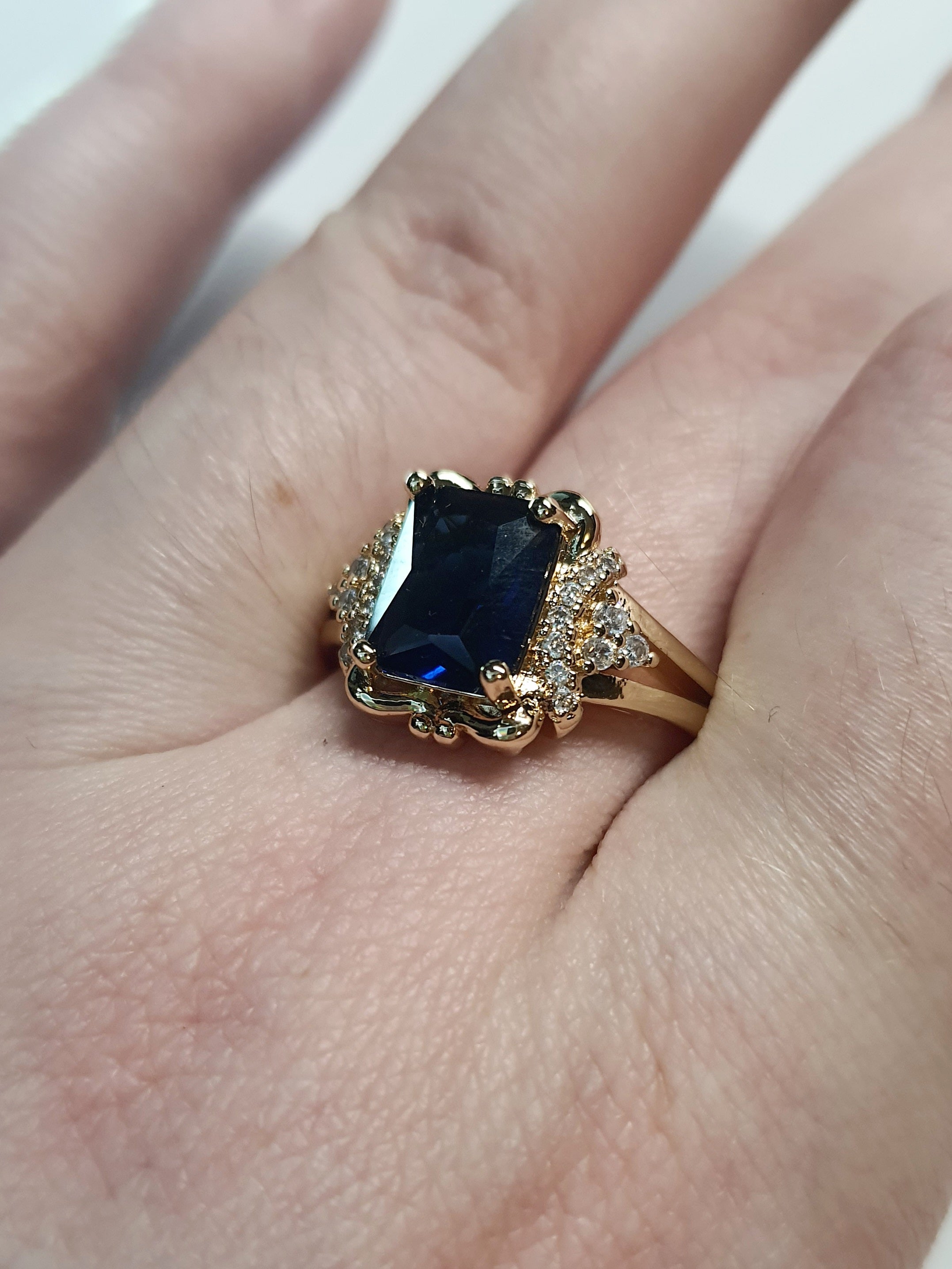 Złoty pierścionek ze stali nierdzewnej z granatowym kamieniem w szlifie szmaragdowym i cyrkoniami po bokach w stylu glamour klasycznym. Rozmiar 20 mm. stal chirurgiczna, stal nierdzewna, wodoodporne, hipoalergiczne, jakość premium, RougeBizu.