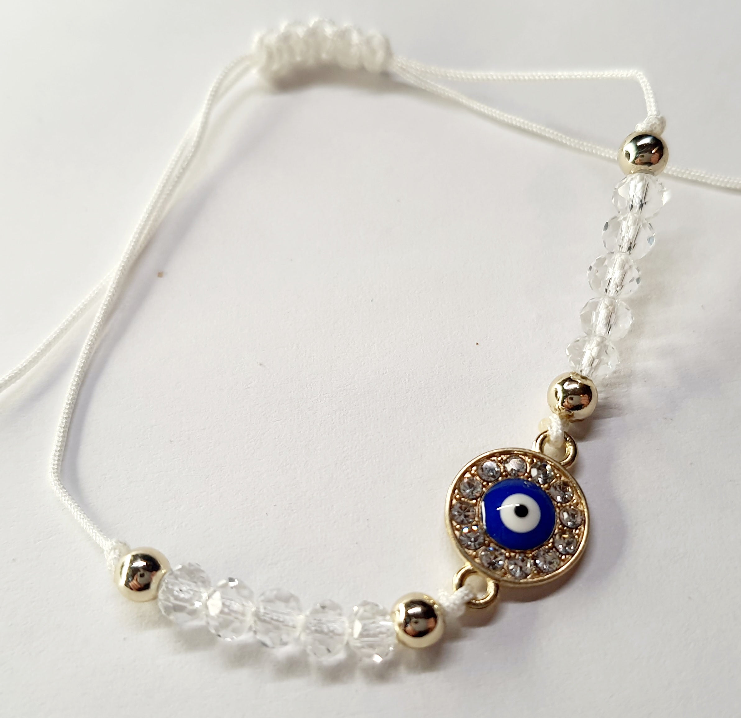 Biała bransoletka na sznurku z niebieskim okiem proroka w złotej oprawie wysadzanej cyrkoniami, z przezroczystymi koralikami. Delikatna, ochronna biżuteria w stylu boho. Wodoodporne, hipoalergiczne, jakość premium. Rouge Bizu.