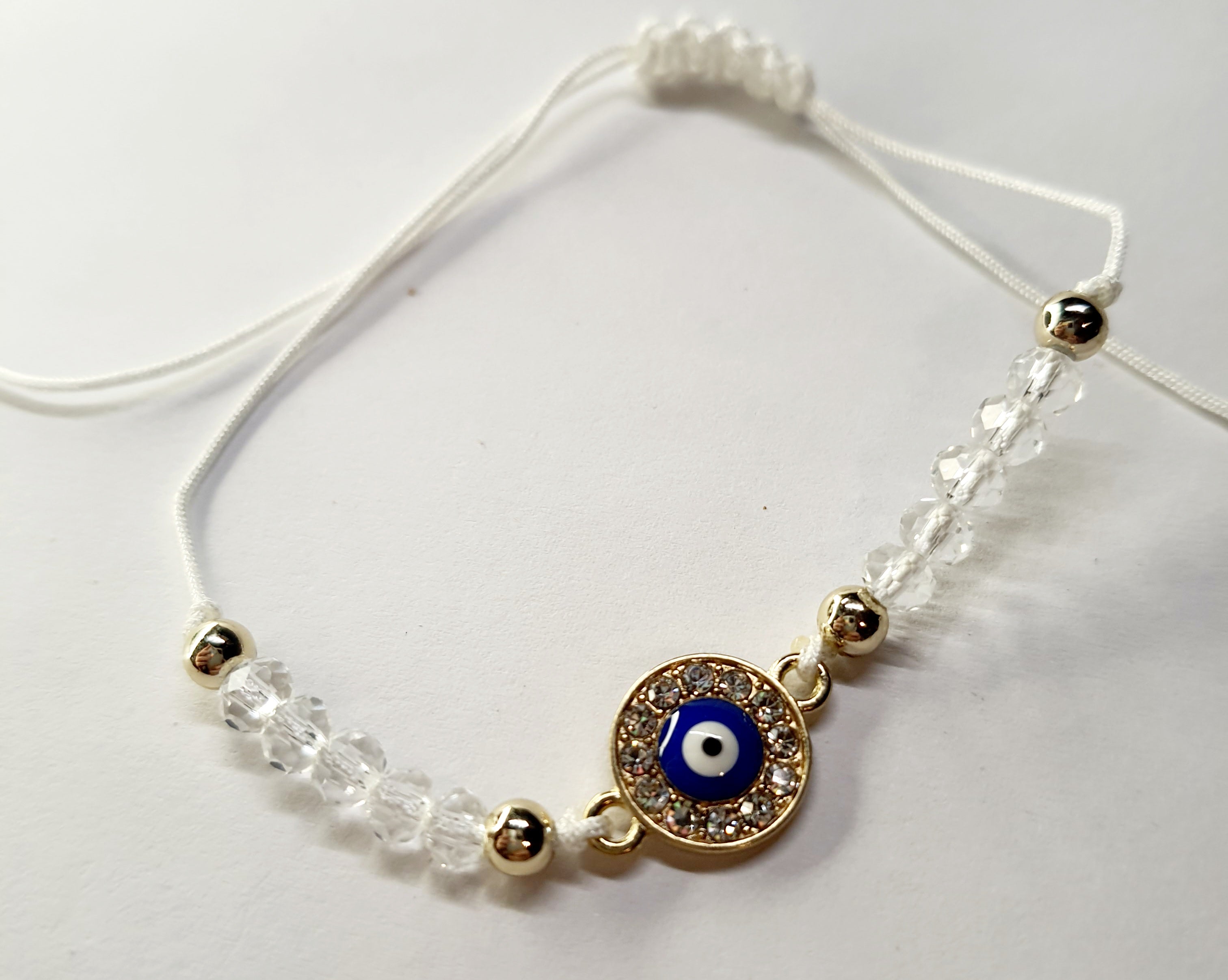Biała bransoletka na sznurku z niebieskim okiem proroka w złotej oprawie wysadzanej cyrkoniami, z przezroczystymi koralikami. Delikatna, ochronna biżuteria w stylu boho. Wodoodporne, hipoalergiczne, jakość premium. Rouge Bizu.