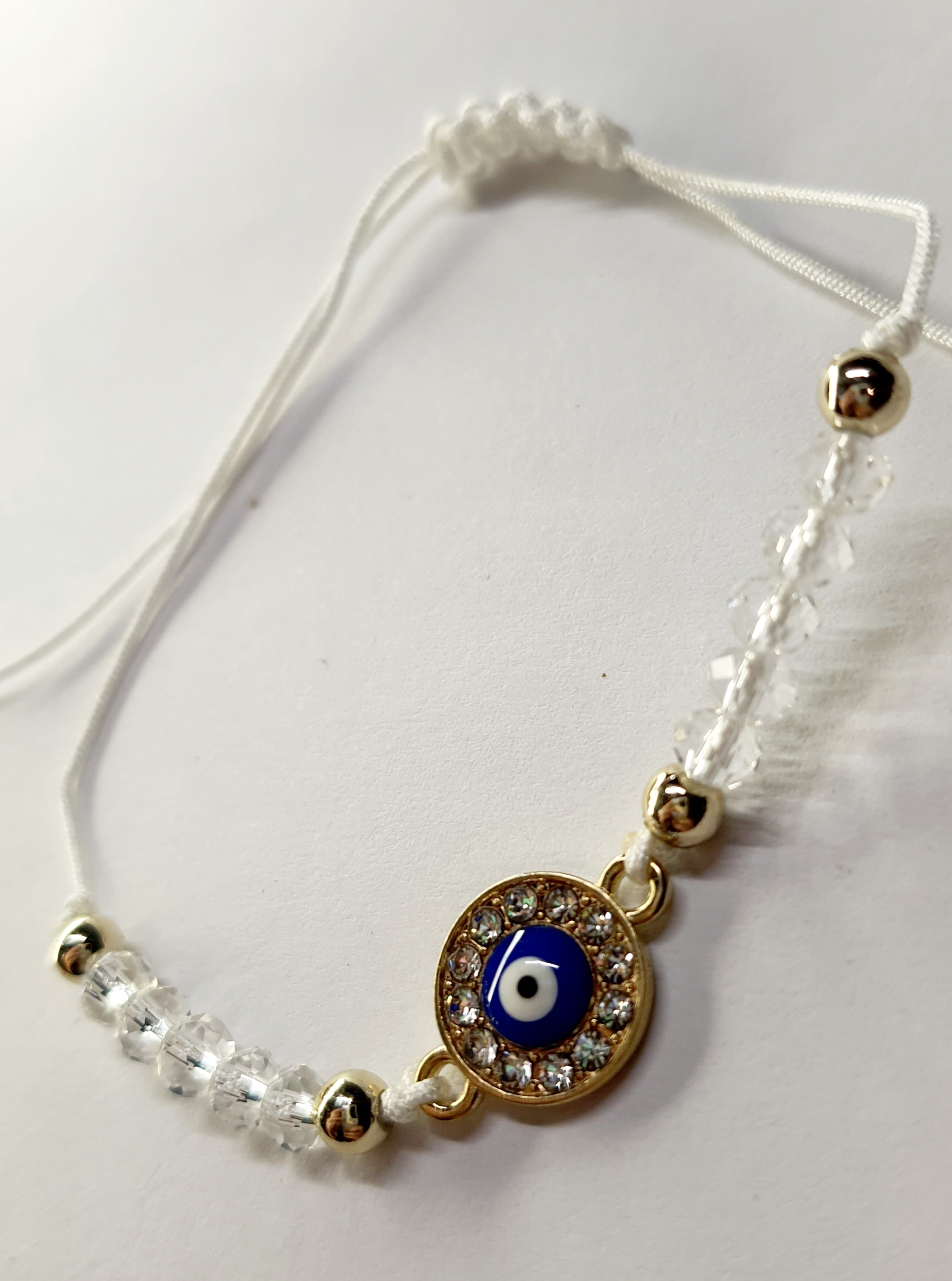 Biała bransoletka na sznurku z niebieskim okiem proroka w złotej oprawie wysadzanej cyrkoniami, z przezroczystymi koralikami. Delikatna, ochronna biżuteria w stylu boho. Wodoodporne, hipoalergiczne, jakość premium. Rouge Bizu.