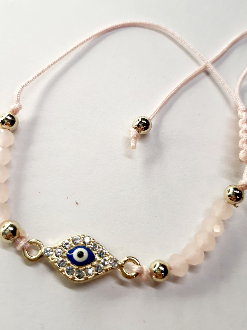 Bransoletka z okiem proroka w złotej oprawie wysadzanej cyrkoniami, na pudroworóżowym sznurku z matowymi różowymi koralikami. Delikatna, kobieca biżuteria ochronna w stylu boho. Wodoodporne, hipoalergiczne, jakość premium. Rouge Bizu.