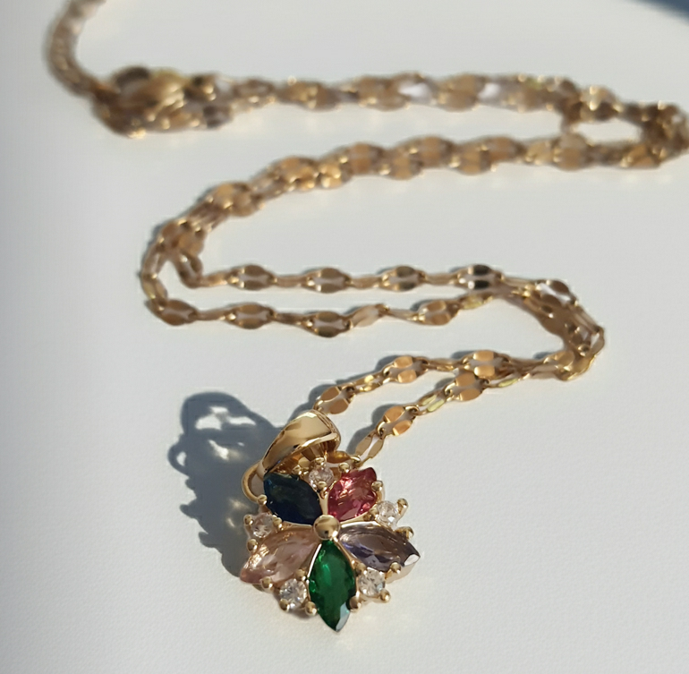 Złoty naszyjnik ze stali chirurgicznej z zawieszką w kształcie kwiatka wysadzanego wielokolorowymi cyrkoniami (zielone, różowe, błękitne, granatowe). Regulowana długość 40–45 cm. Stal chirurgiczna, stal nierdzewna, wodoodporne, hipoalergiczne, jakość premium. RougeBizu.