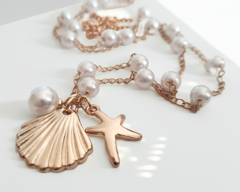 Naszyjnik z drobnymi syntetycznymi perełkami i złotymi zawieszkami w kształcie muszelki i rozgwiazdy. Długość regulowana 55–56 cm. Wykonany ze stali nierdzewnej (chirurgicznej). Delikatna biżuteria w stylu morskim. Rouge Bizu.
