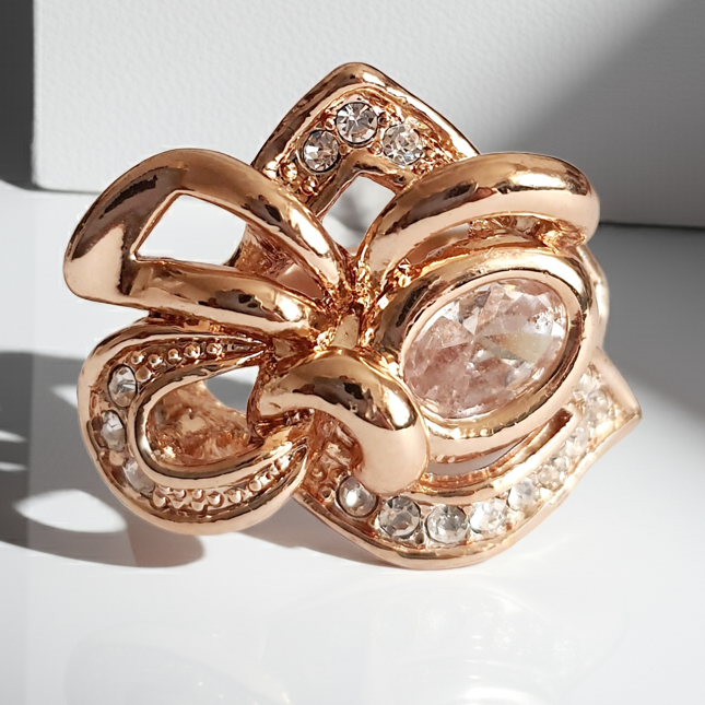 Pierścionek ze stali chirurgicznej w kolorze żółtego złota, z owalnym bezbarwnym kryształem i wijącymi się ramionami zdobionymi cyrkoniami. Styl glamour. Rozmiar 17 mm. Rouge Bizu.
