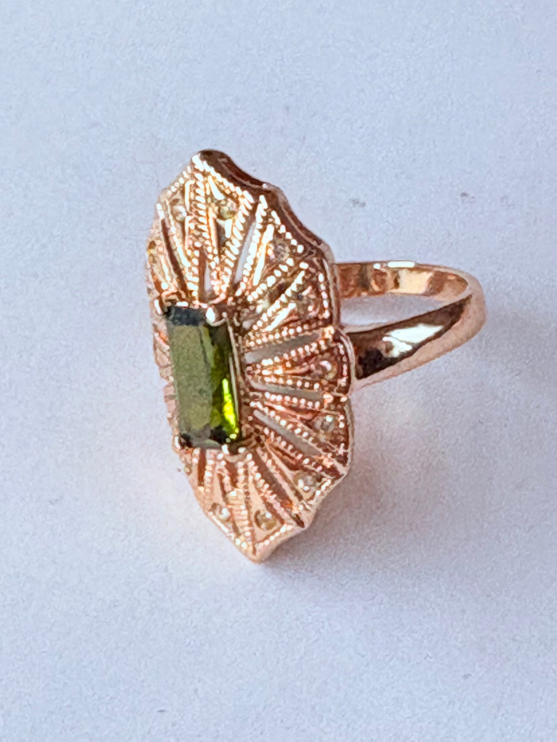 Pierścionek z podłużnym szmaragdowym kamieniem w stylu glamour vintage, oprawiony w wachlarz różowozłotych detali z drobnymi cyrkoniami. Rozmiar 20 mm. Elegancki, kobiecy, wykonany ze stali chirurgicznej. Rouge Bizu.

