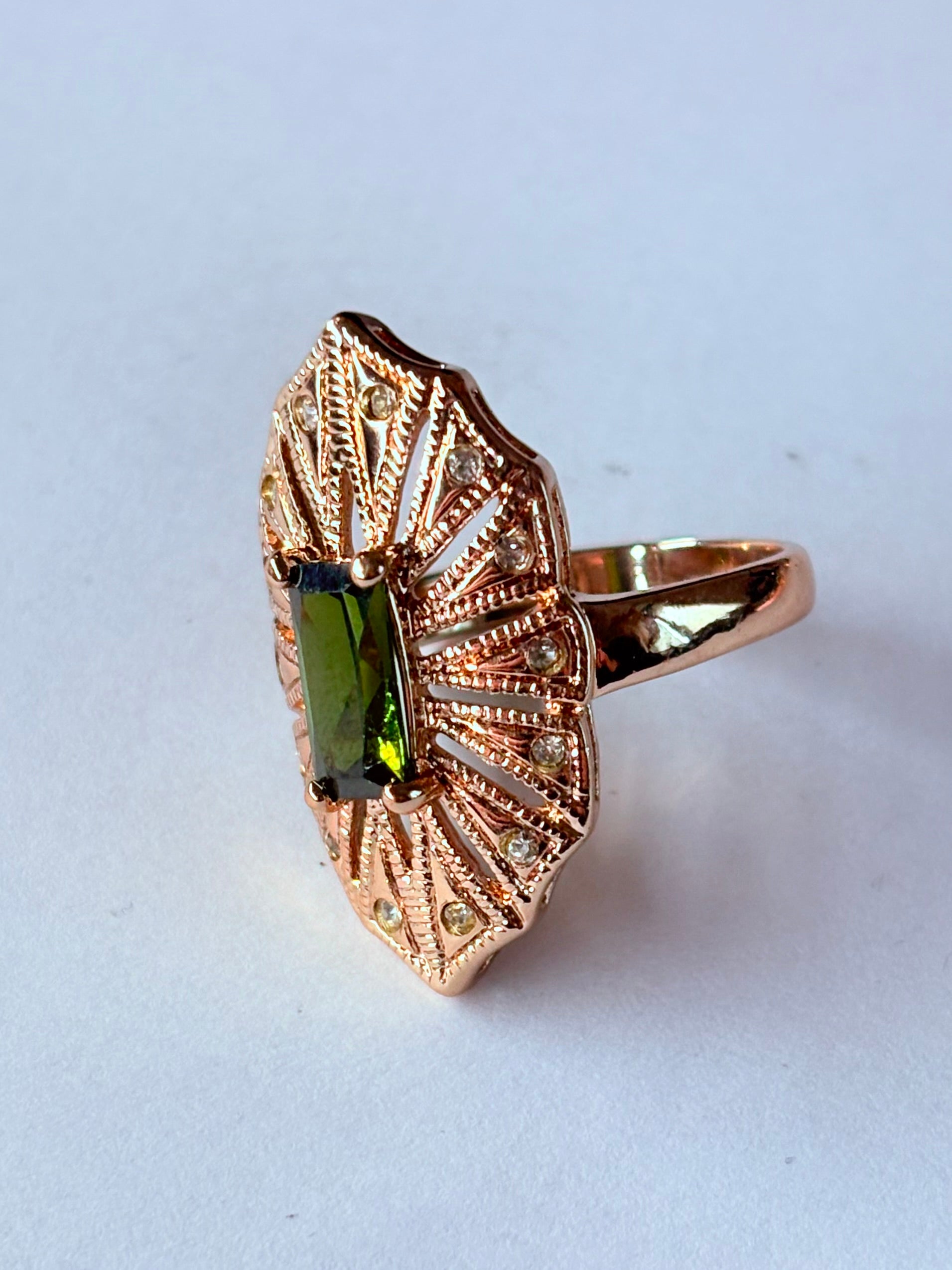 Pierścionek z podłużnym szmaragdowym kamieniem w stylu glamour vintage, oprawiony w wachlarz różowozłotych detali z drobnymi cyrkoniami. Rozmiar 20 mm. Elegancki, kobiecy, wykonany ze stali chirurgicznej. Rouge Bizu.


