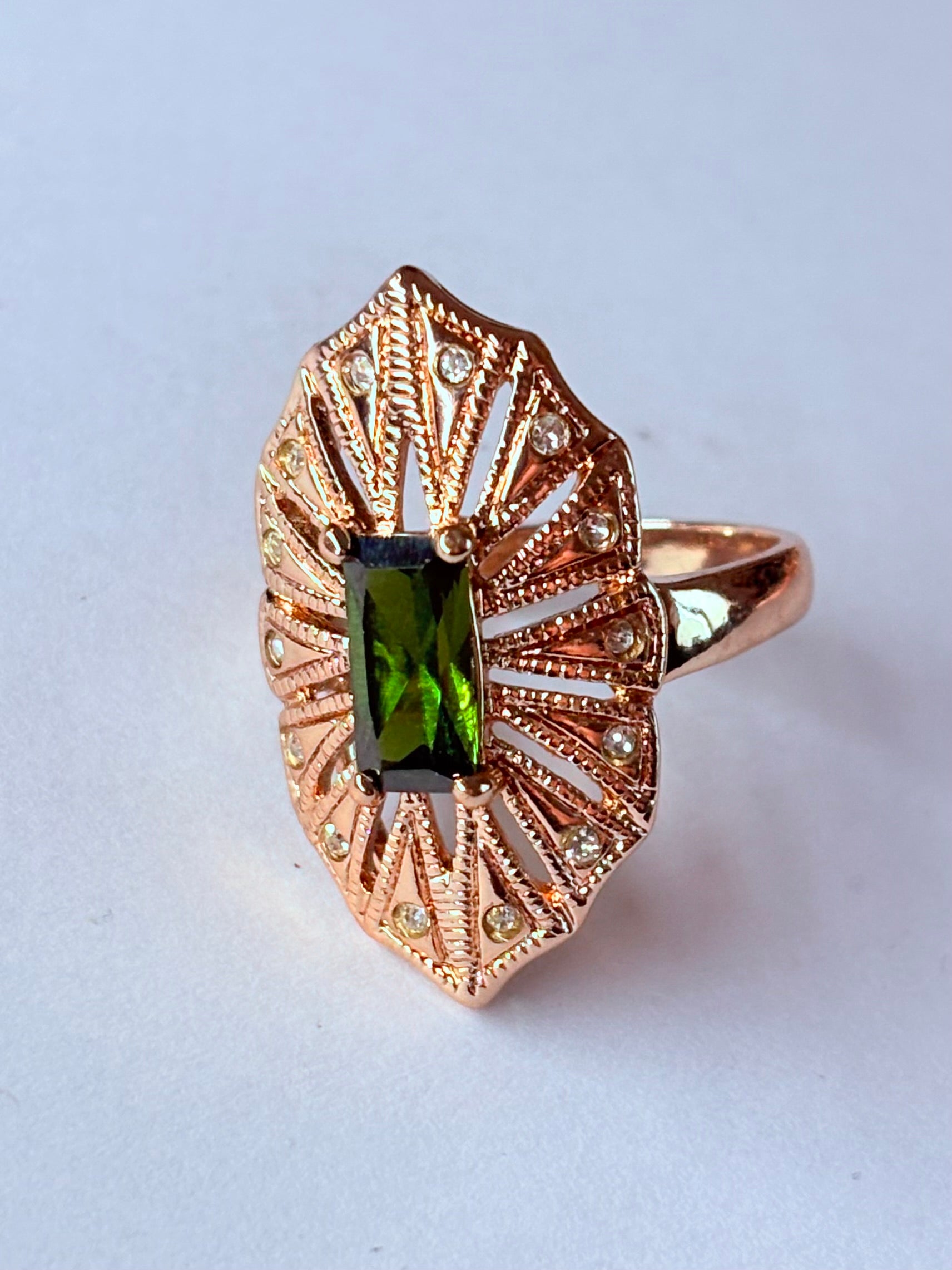 Pierścionek z podłużnym szmaragdowym kamieniem w stylu glamour vintage, oprawiony w wachlarz różowozłotych detali z drobnymi cyrkoniami. Rozmiar 20 mm. Elegancki, kobiecy, wykonany ze stali chirurgicznej. Rouge Bizu.

