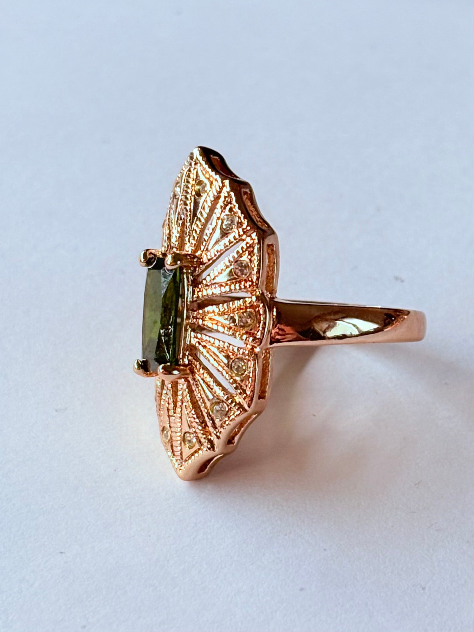 Pierścionek z podłużnym szmaragdowym kamieniem w stylu glamour vintage, oprawiony w wachlarz różowozłotych detali z drobnymi cyrkoniami. Rozmiar 20 mm. Elegancki, kobiecy, wykonany ze stali chirurgicznej. Rouge Bizu.

