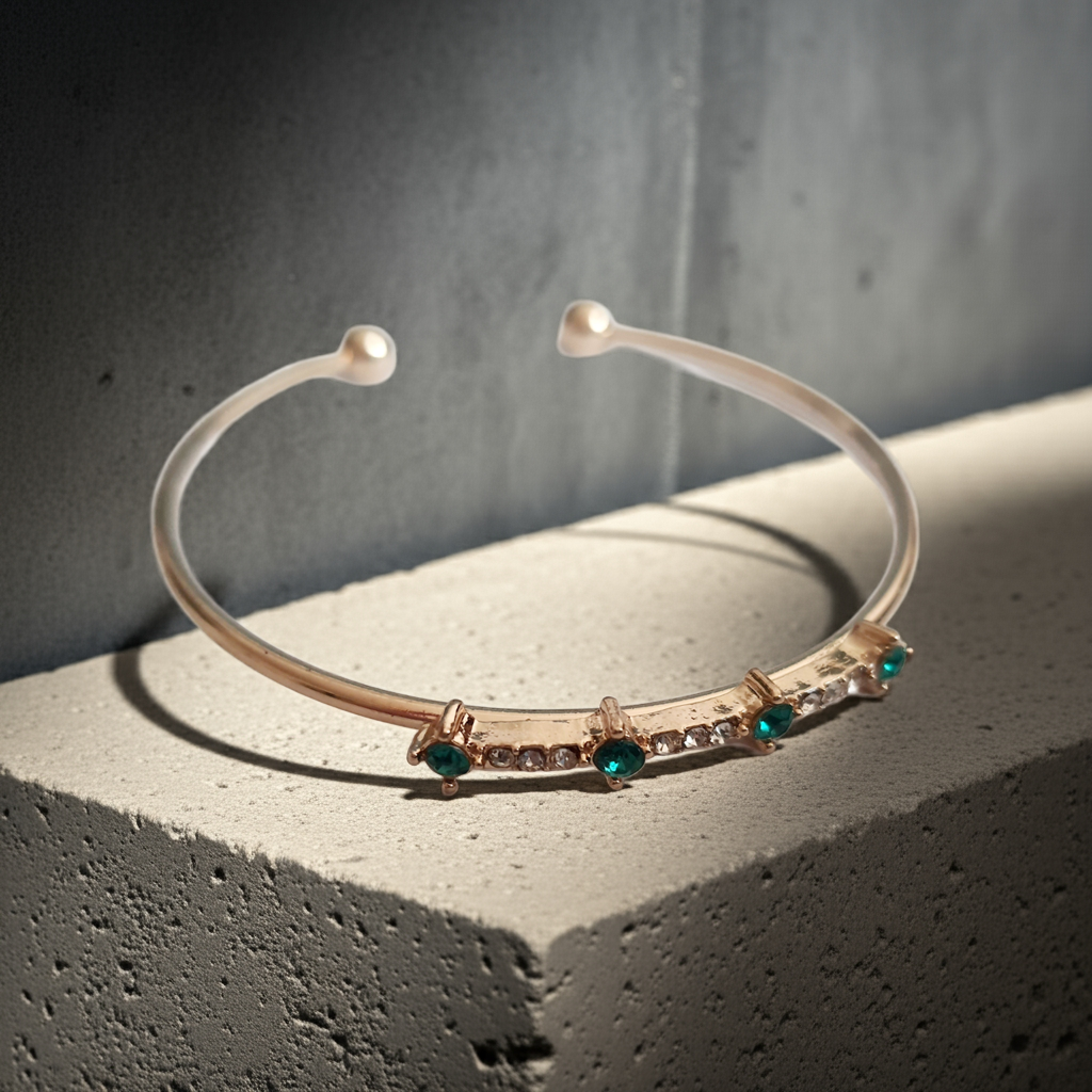 Sztywna złota bransoletka damska typu bangle ze stali nierdzewnej, ozdobiona szmaragdowymi kamieniami i drobnymi cyrkoniami. Styl glamour, elegancja i wyrazistość. Rouge Bizu.

