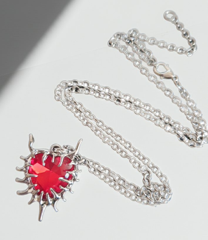 Naszyjnik z czerwonym kamieniem w kształcie serca, otoczonym srebrzystą oprawą przypominającą płomienie. Styl gotycki i wyrazisty. Długość łańcuszka 61 cm. Stal chirurgiczna, stal nierdzewna, wodoodporne, hipoalergiczne, jakość premium. RougeBizu.