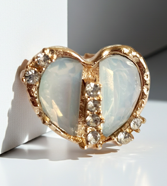 Romantyczny pierścionek ze stali nierdzewnej z podwójnym sercem z opalizującym, mlecznobiałym kamieniem i rzędem błyszczących cyrkonii. Styl glamour, rozmiar 17 mm, regulowany. Rouge Bizu.
