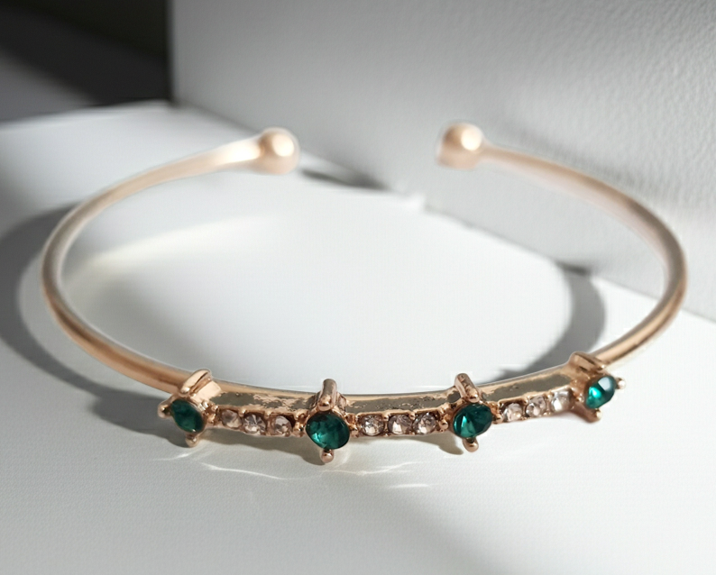 Sztywna złota bransoletka damska typu bangle ze stali nierdzewnej, ozdobiona szmaragdowymi kamieniami i drobnymi cyrkoniami. Styl glamour, elegancja i wyrazistość. Rouge Bizu.
